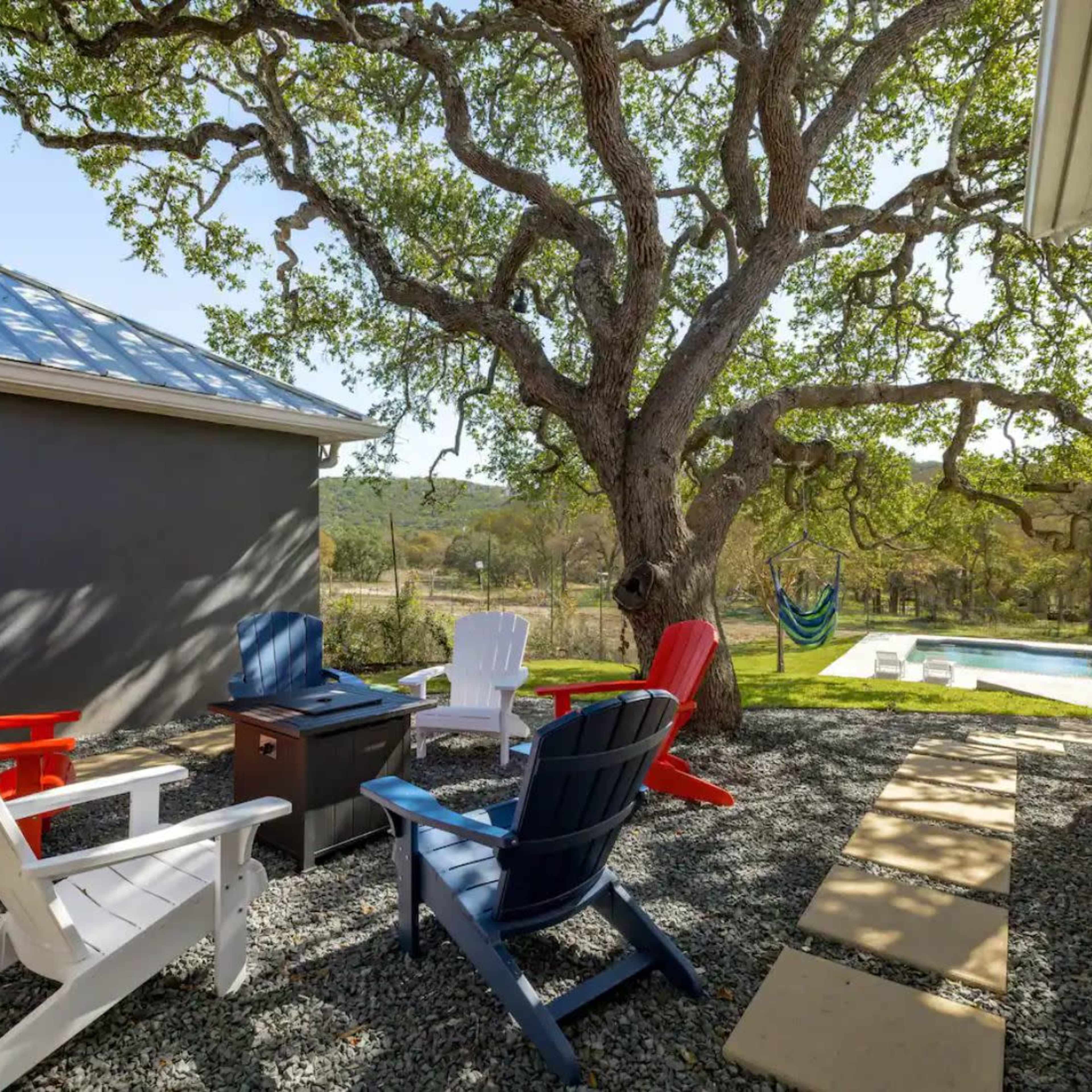 A seating area with colorful Adirondack chairs surrounds a small table beside a tree, with a view of a pool in the background.