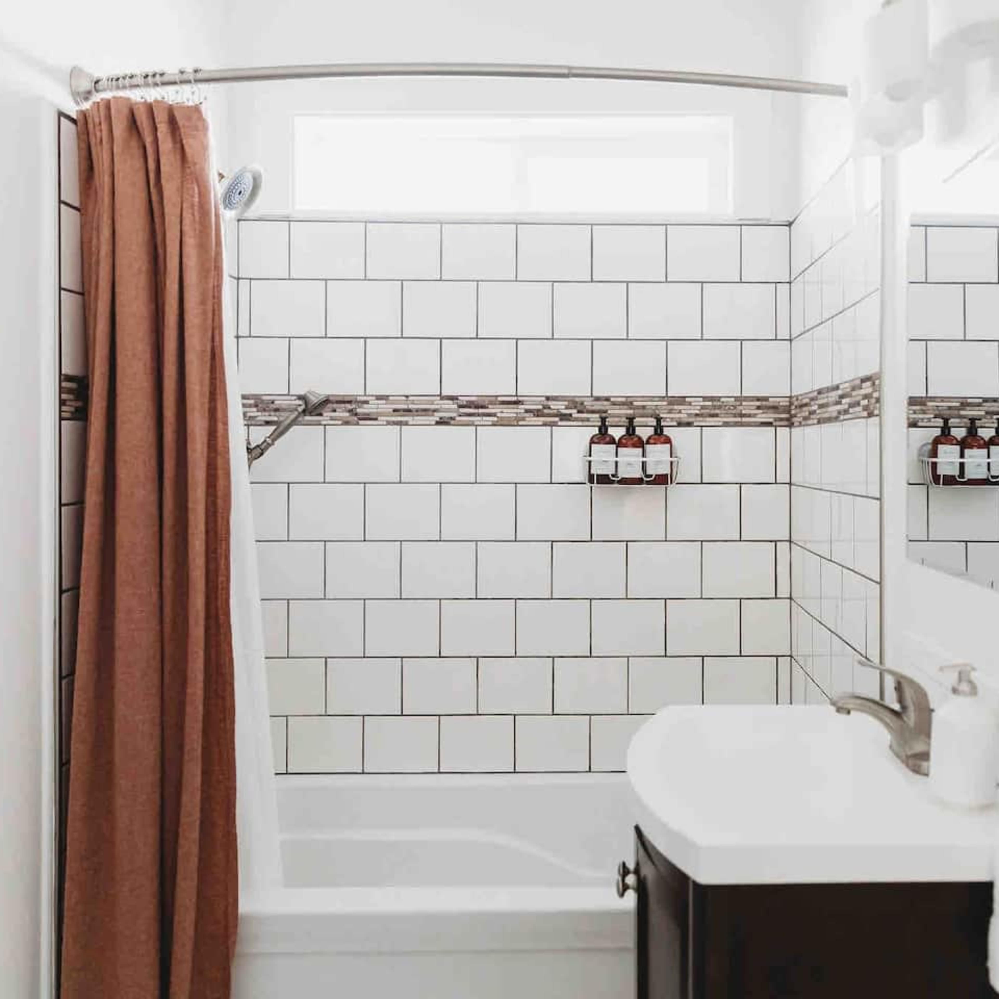 A bathroom featuring a bathtub with a shower, white tiled walls, a brown shower curtain, and a small sink with toiletries displayed on the wall.
