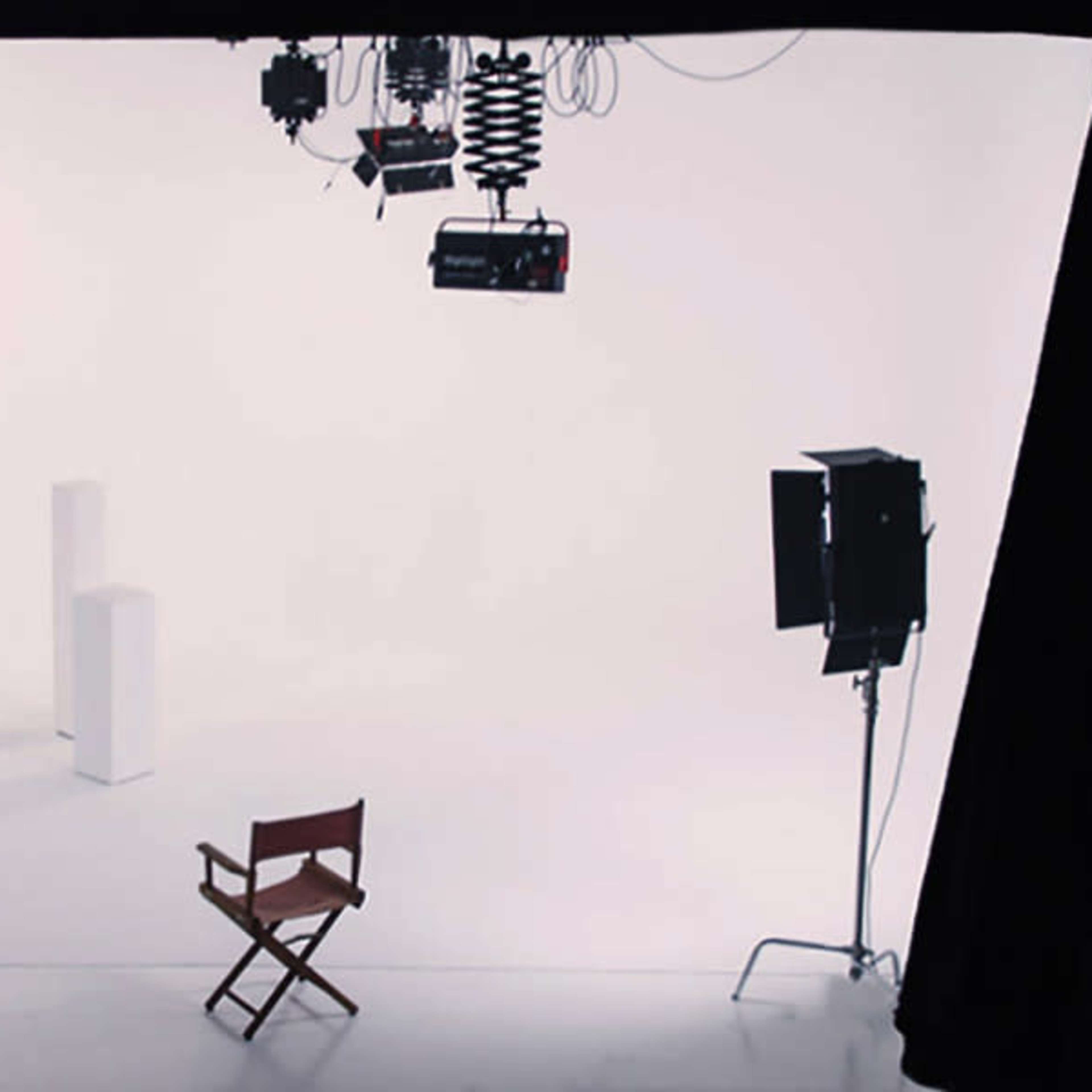 A white studio backdrop with two light stands, two light sources, and a wooden director's chair placed in front of two white pedestals.