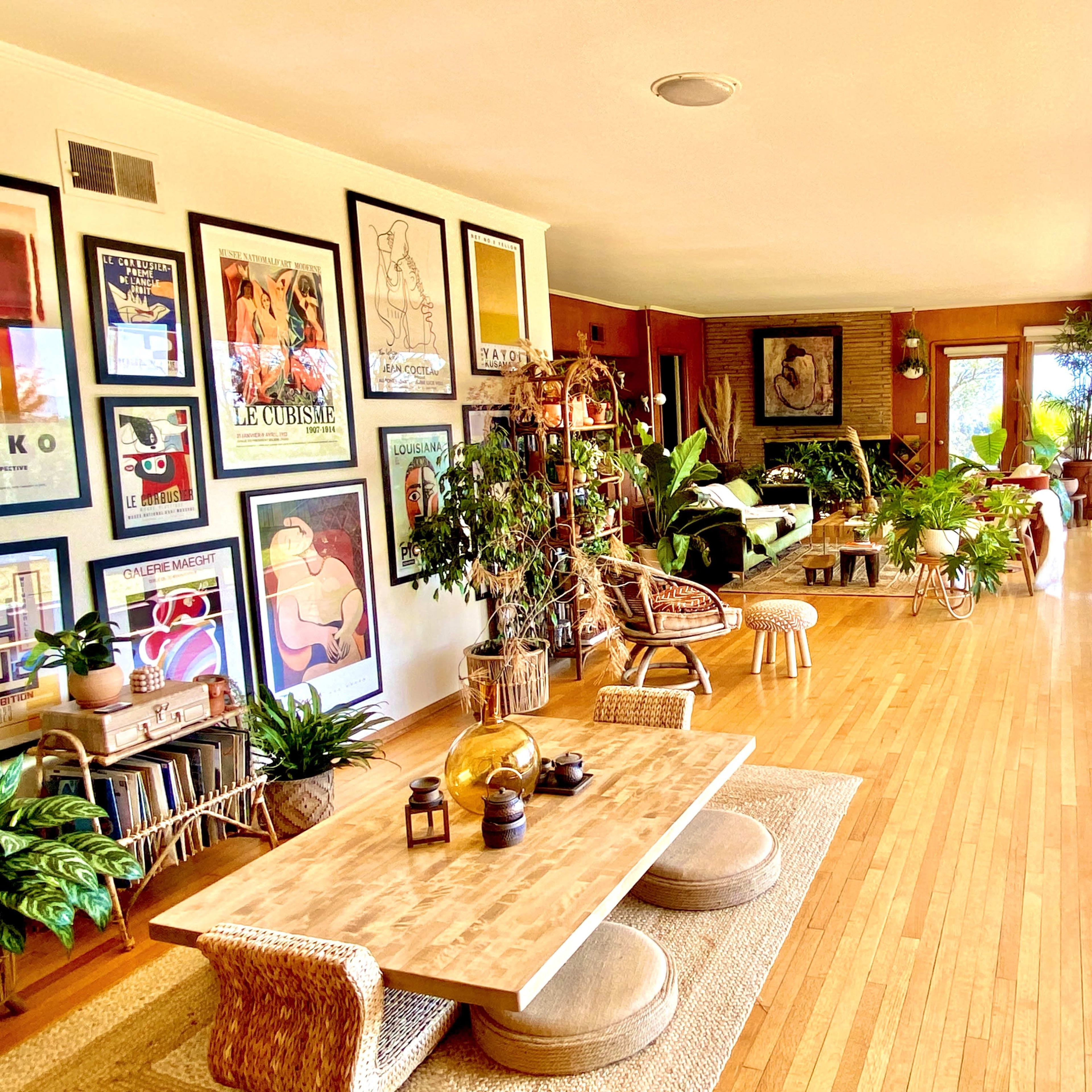 A spacious living area adorned with framed artwork, wooden furniture, and numerous plants, with large windows that open to a view of the outdoors.