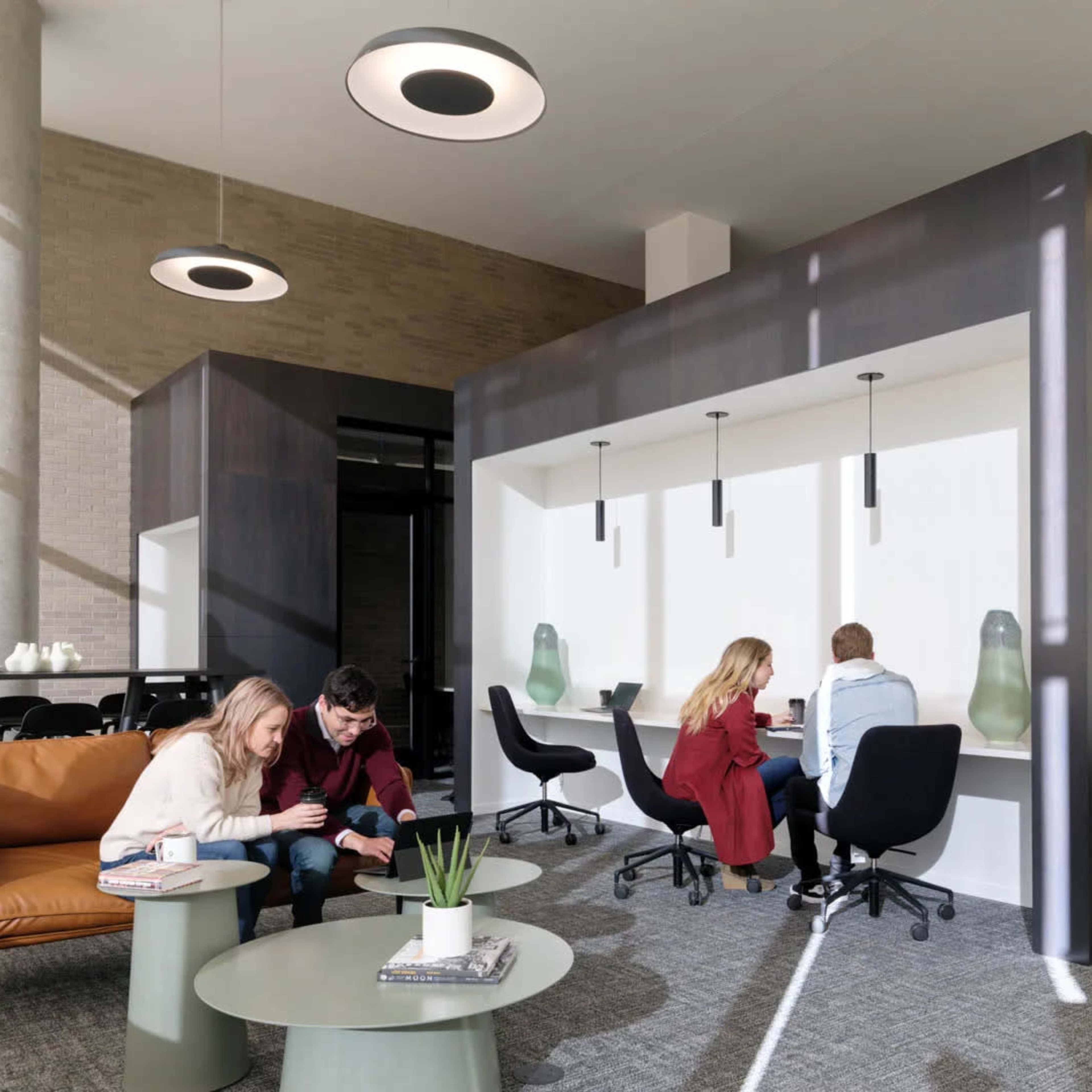 A modern office space features two groups of people working at tables, with a lounge area and private booths for collaboration.
