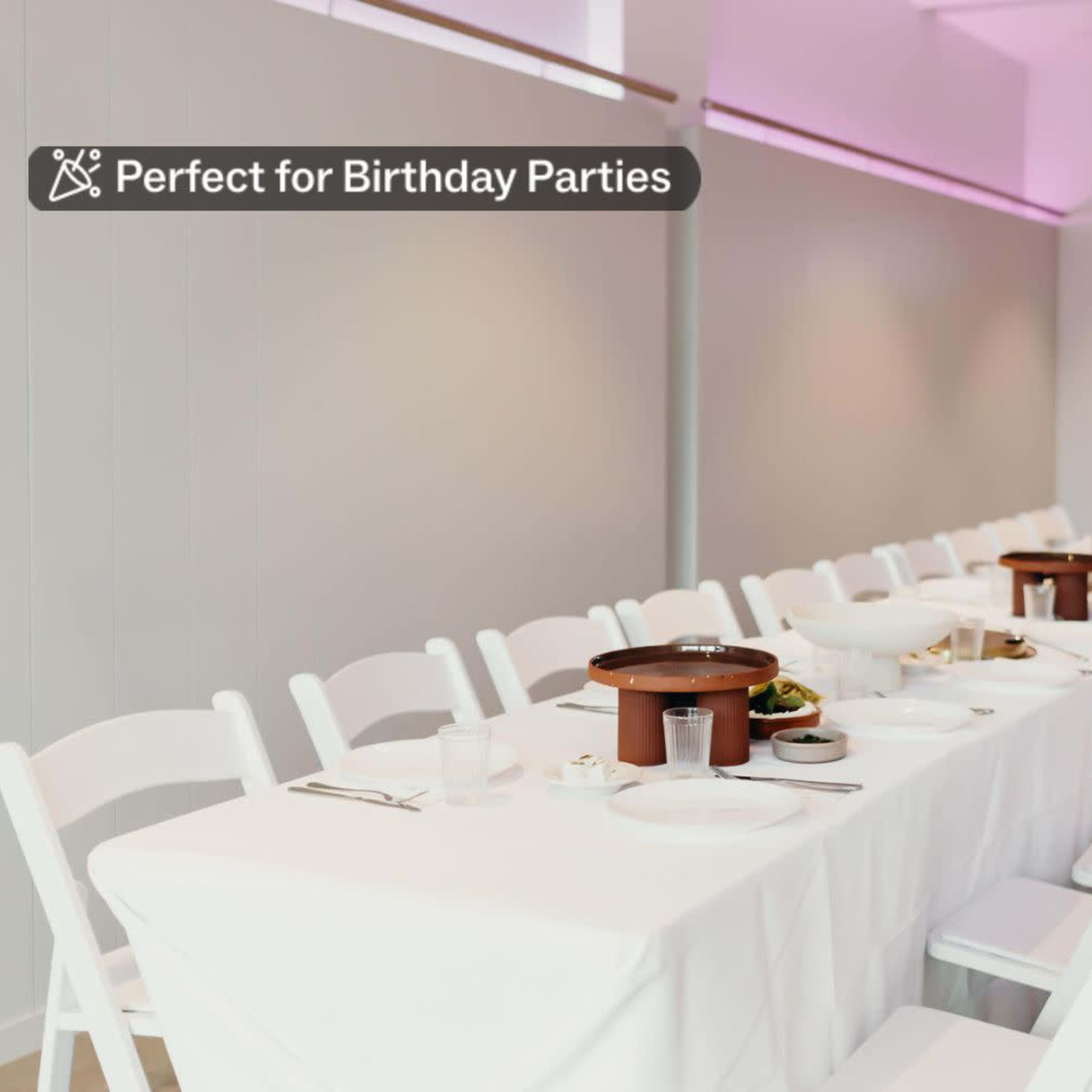 A long, white table is set for a birthday party with plates and bowls, and a neon sign that reads "Baba" on the wall.