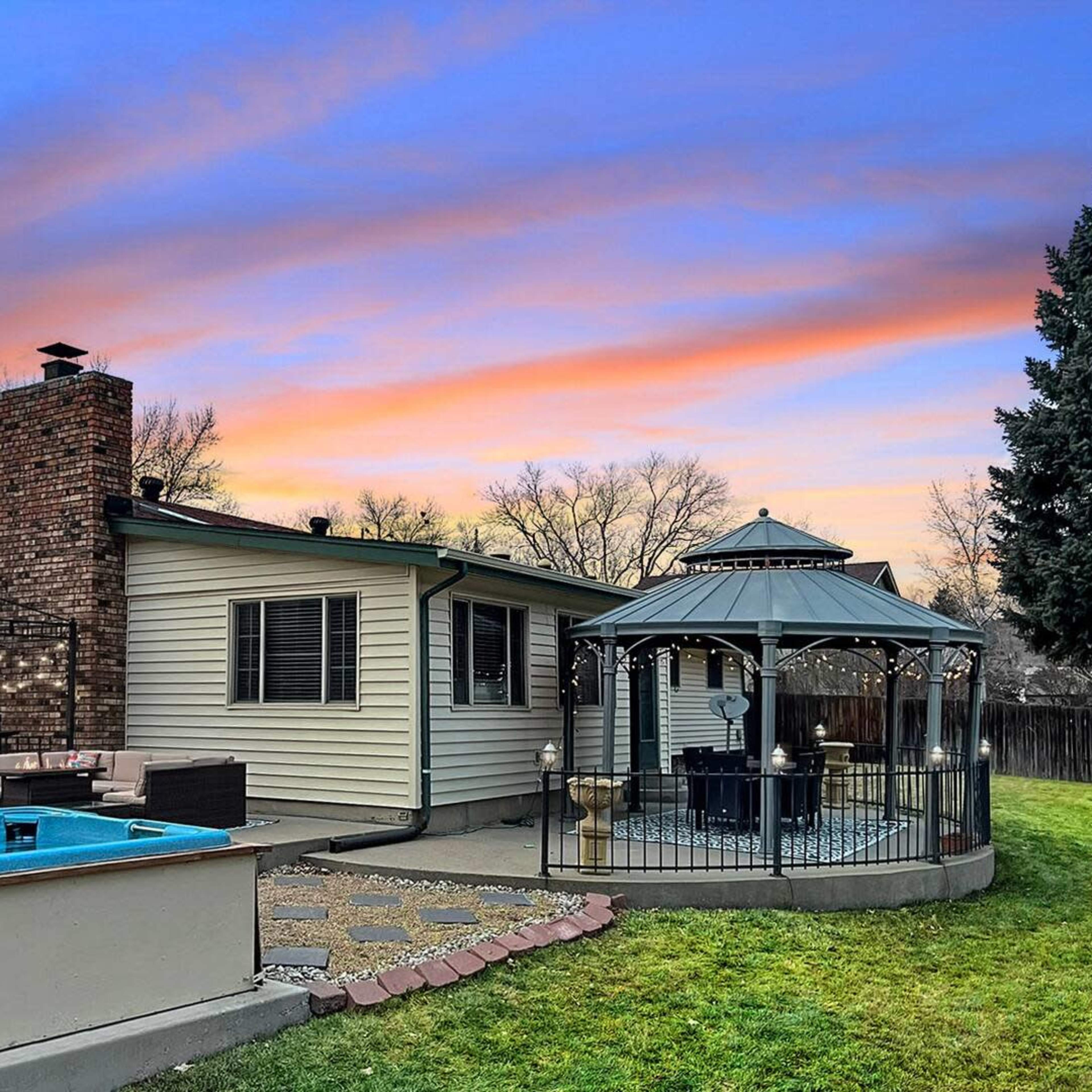 A backyard scene features a rectangular pool, a gazebo, and a house with a brick chimney against a colorful sunset sky.