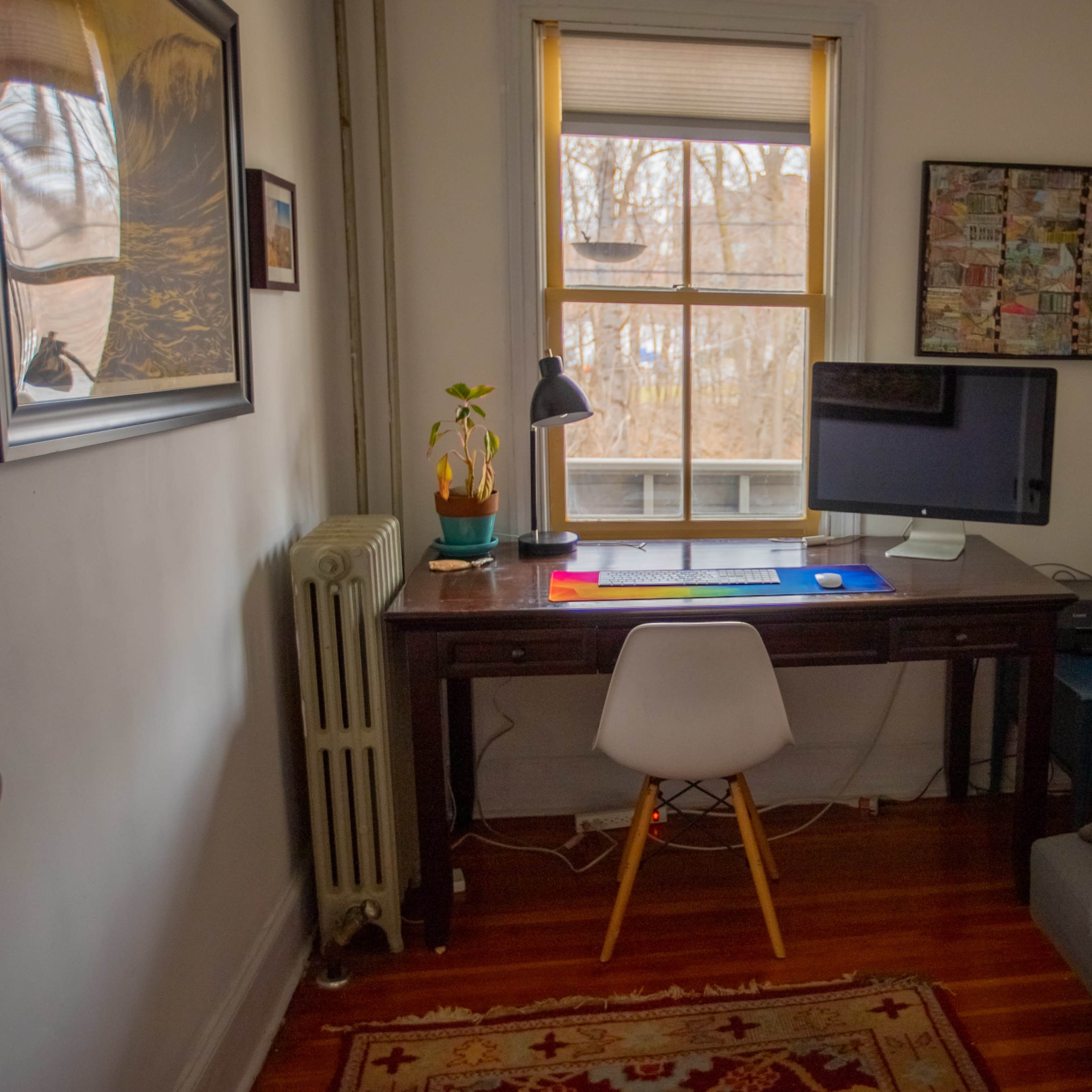A small office space features a wooden desk with a computer and lamp, a gray sofa, and a window overlooking a wooded area.