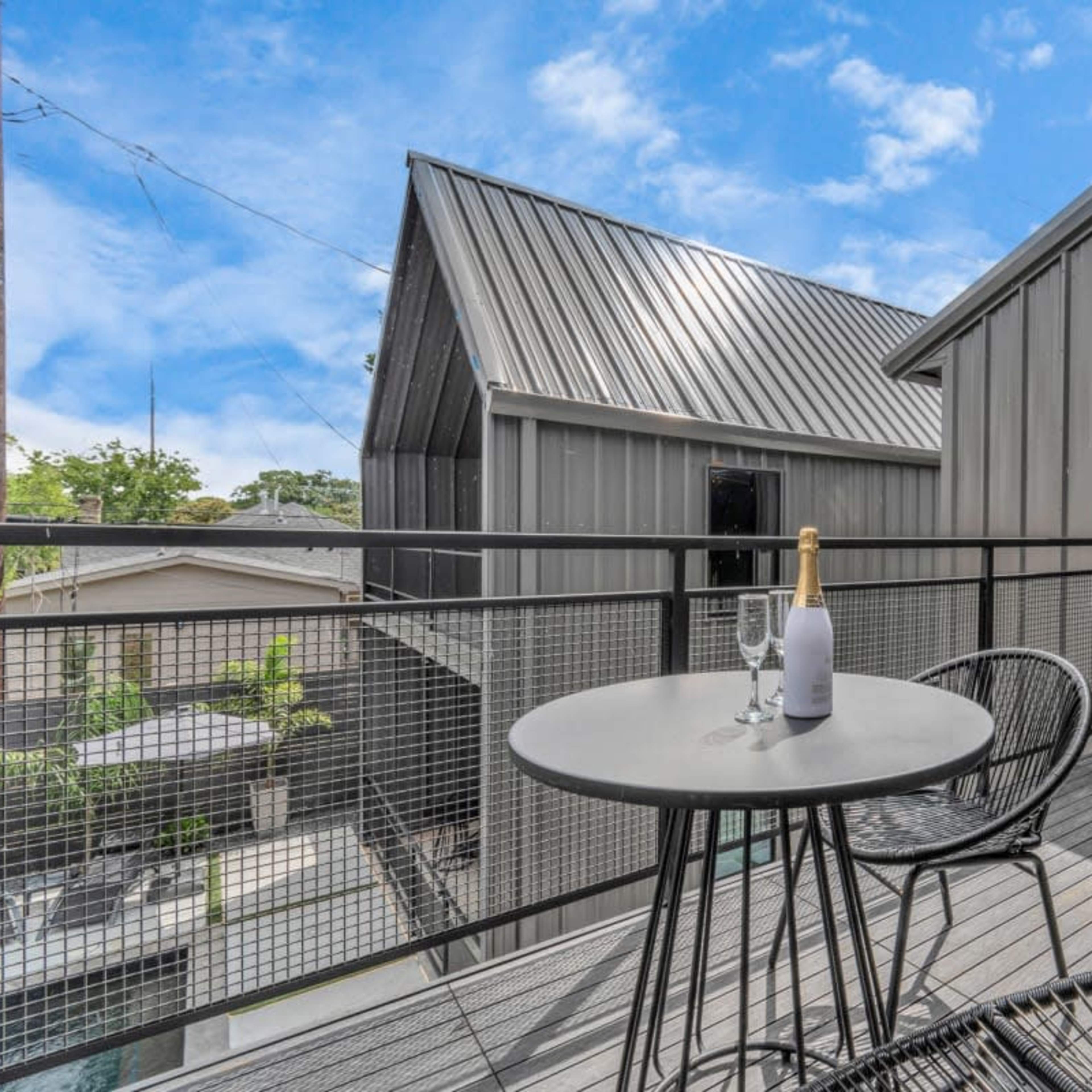 A small outdoor balcony features a table set with a champagne bottle and glasses, overlooking buildings with metal roofs under a blue sky.