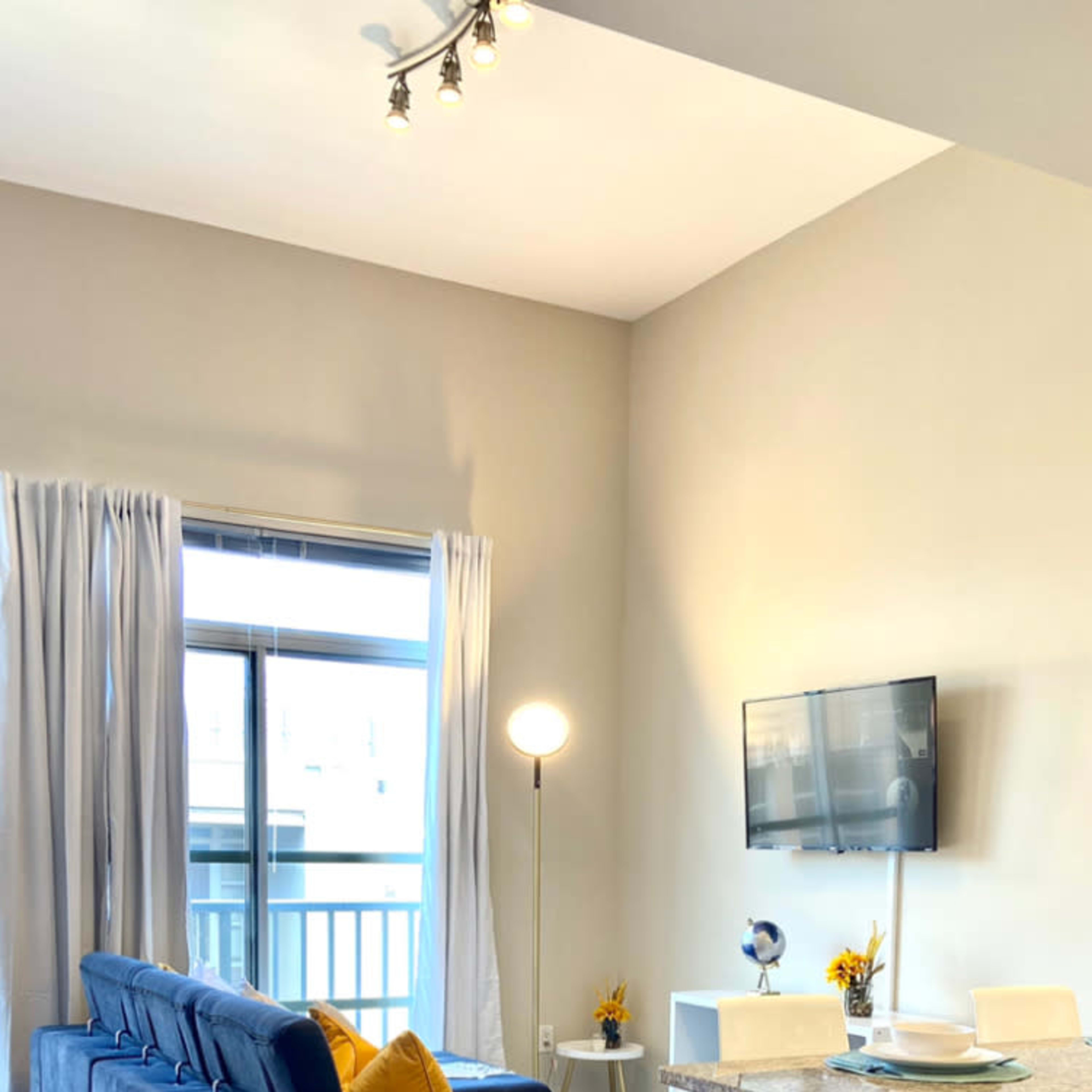 A modern living area with a blue sofa, a small dining table, and a TV mounted on the wall, all illuminated by natural light from large windows.