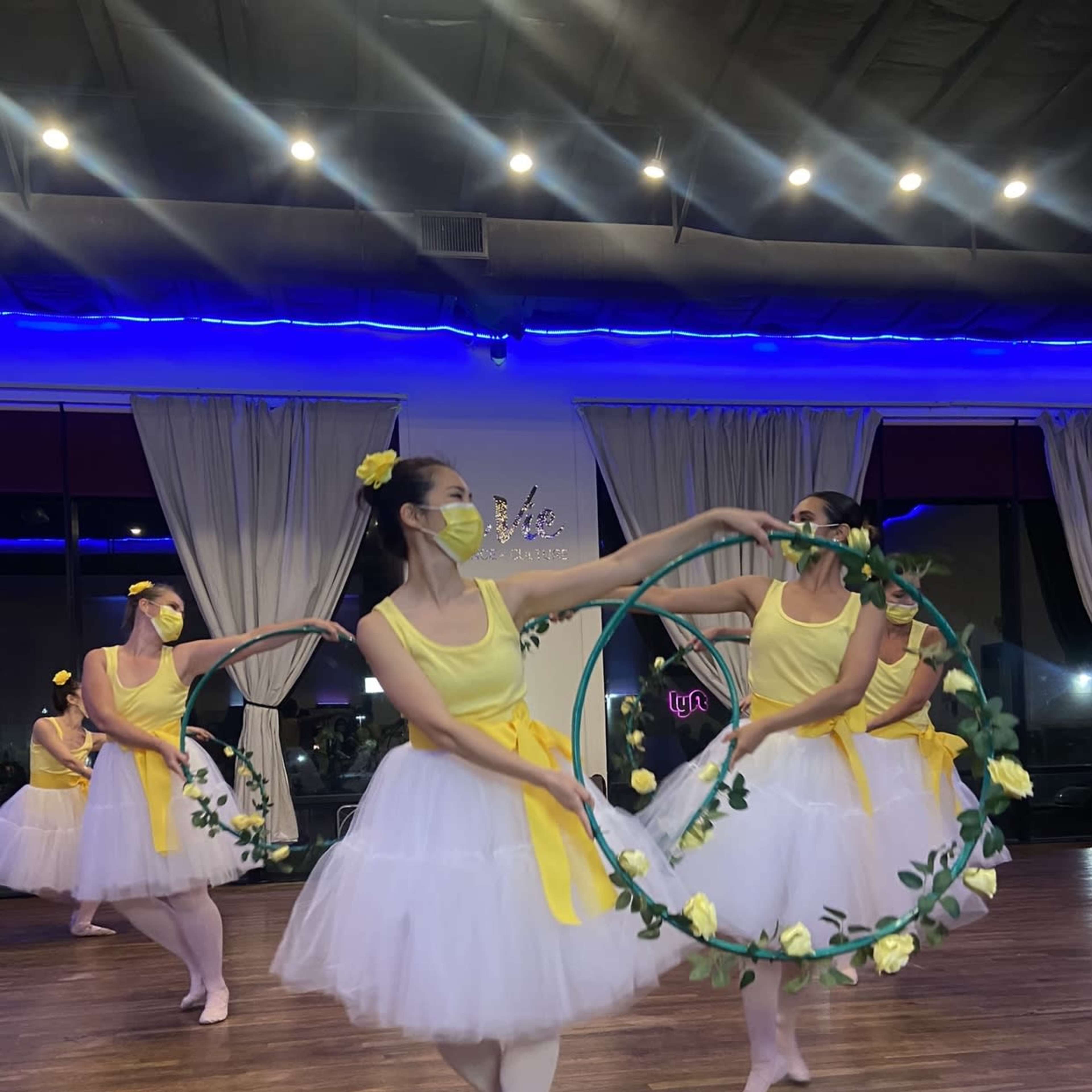 A group of dancers in yellow tops and white tutus perform with hoops adorned with flowers in a brightly lit studio.