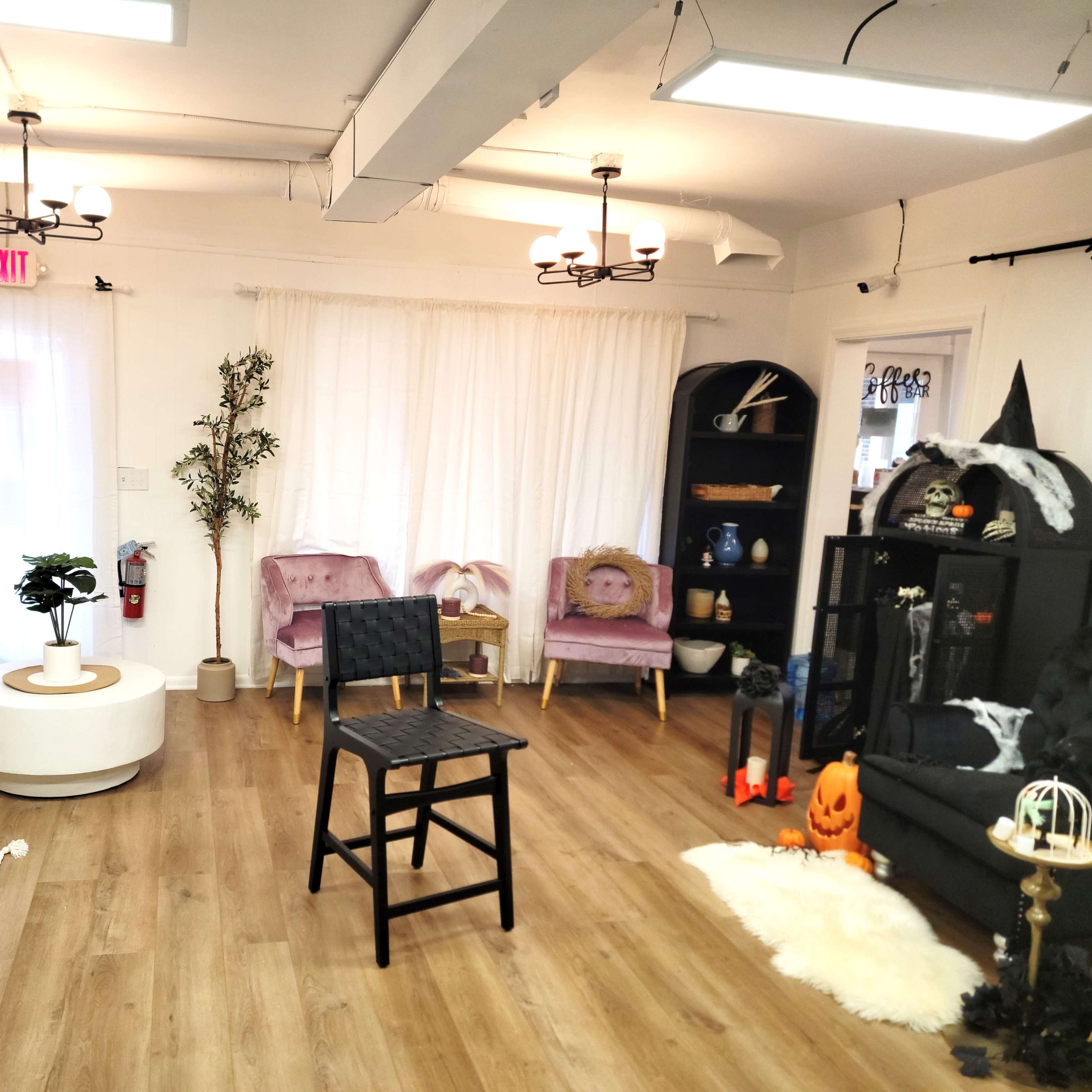 The room features light-colored walls, wooden flooring, and a variety of furniture, including a black chair, pink armchairs, and a decorative bookshelf.