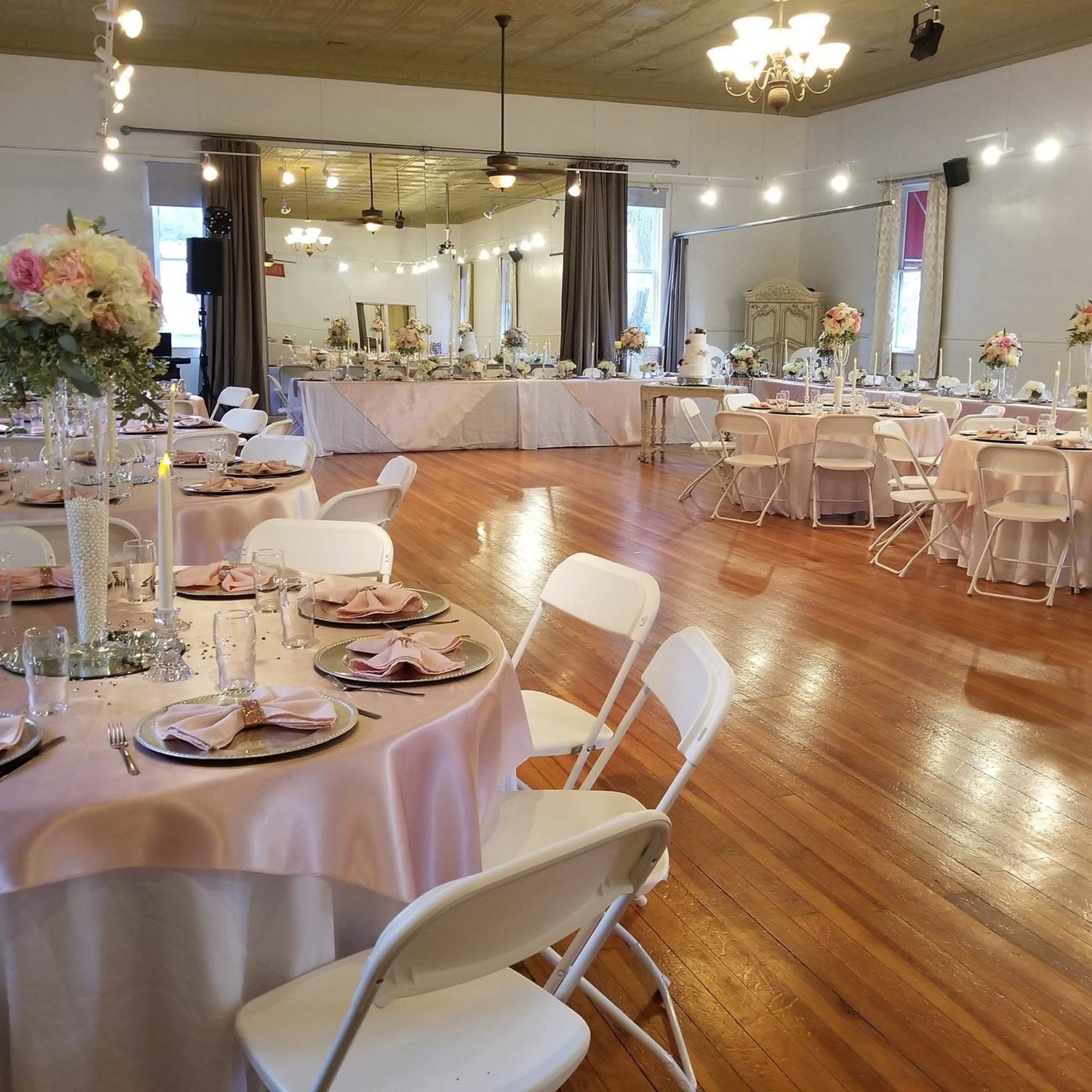 A banquet hall set up for an event, featuring round tables with pink tablecloths and white chairs, adorned with floral centerpieces and candles, alongside a buffet table in the background.