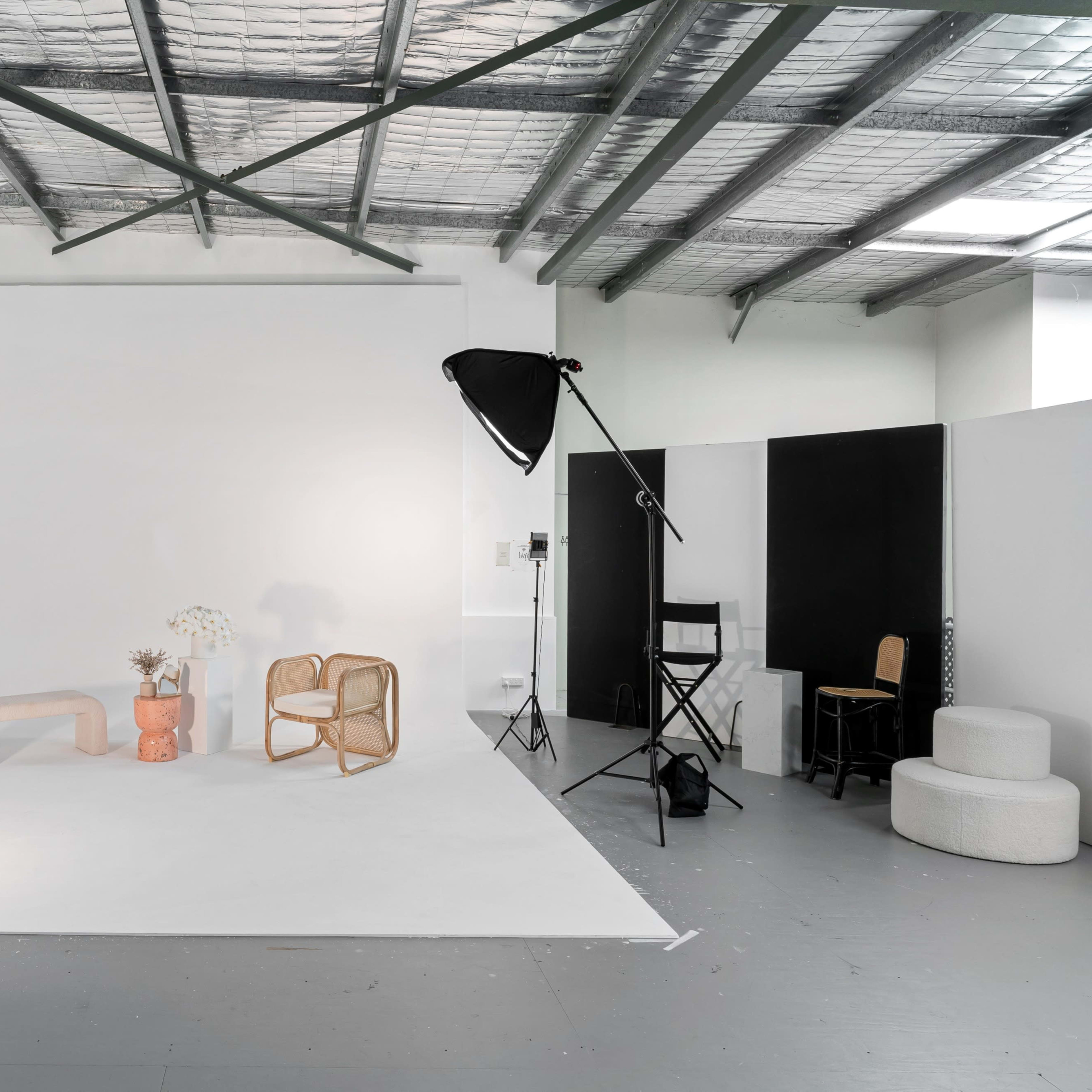 A brightly lit photo studio featuring a white backdrop, various furniture pieces, and lighting equipment.