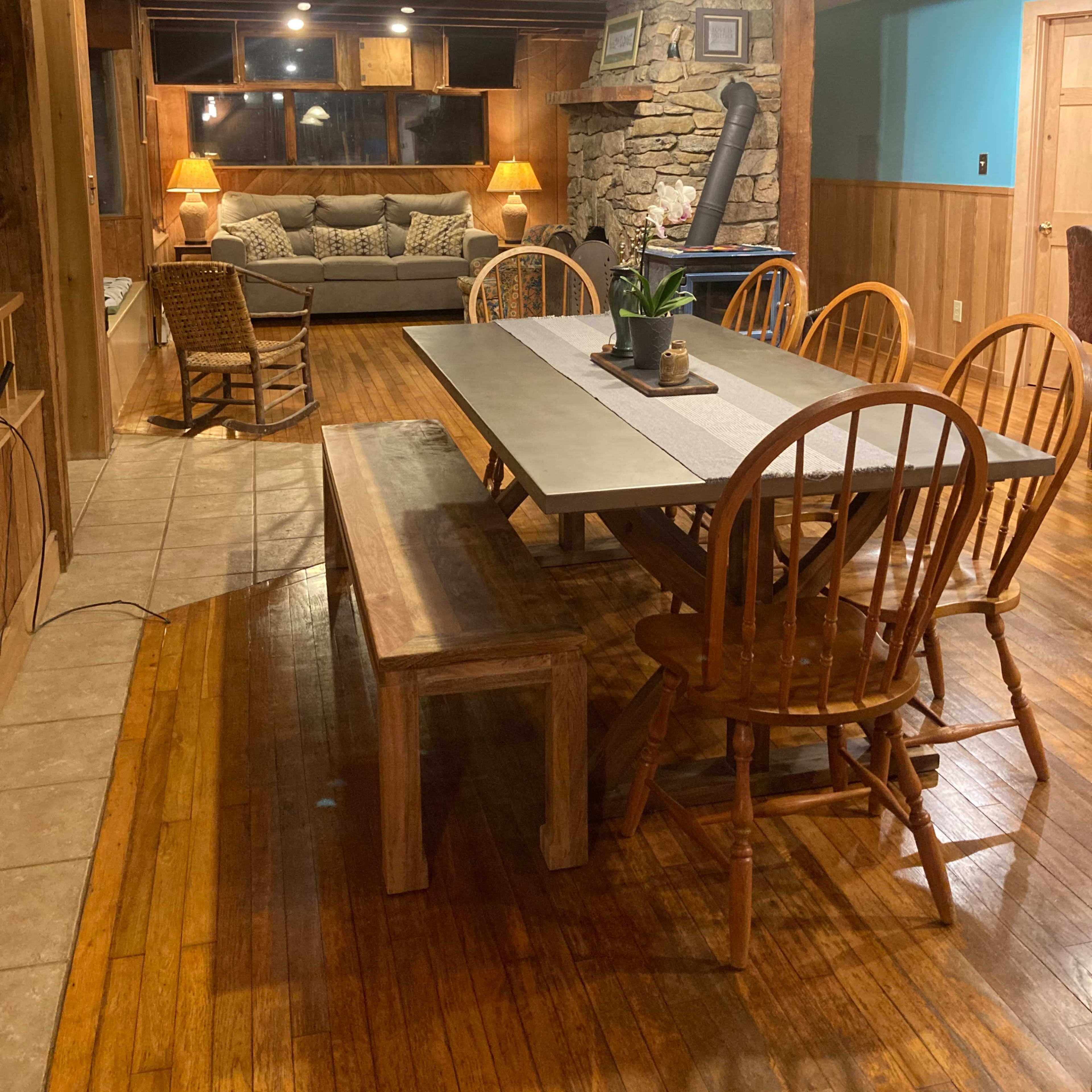 A wooden dining area with a long table and chairs, adjacent to a cozy living space featuring a couch and a stone fireplace.