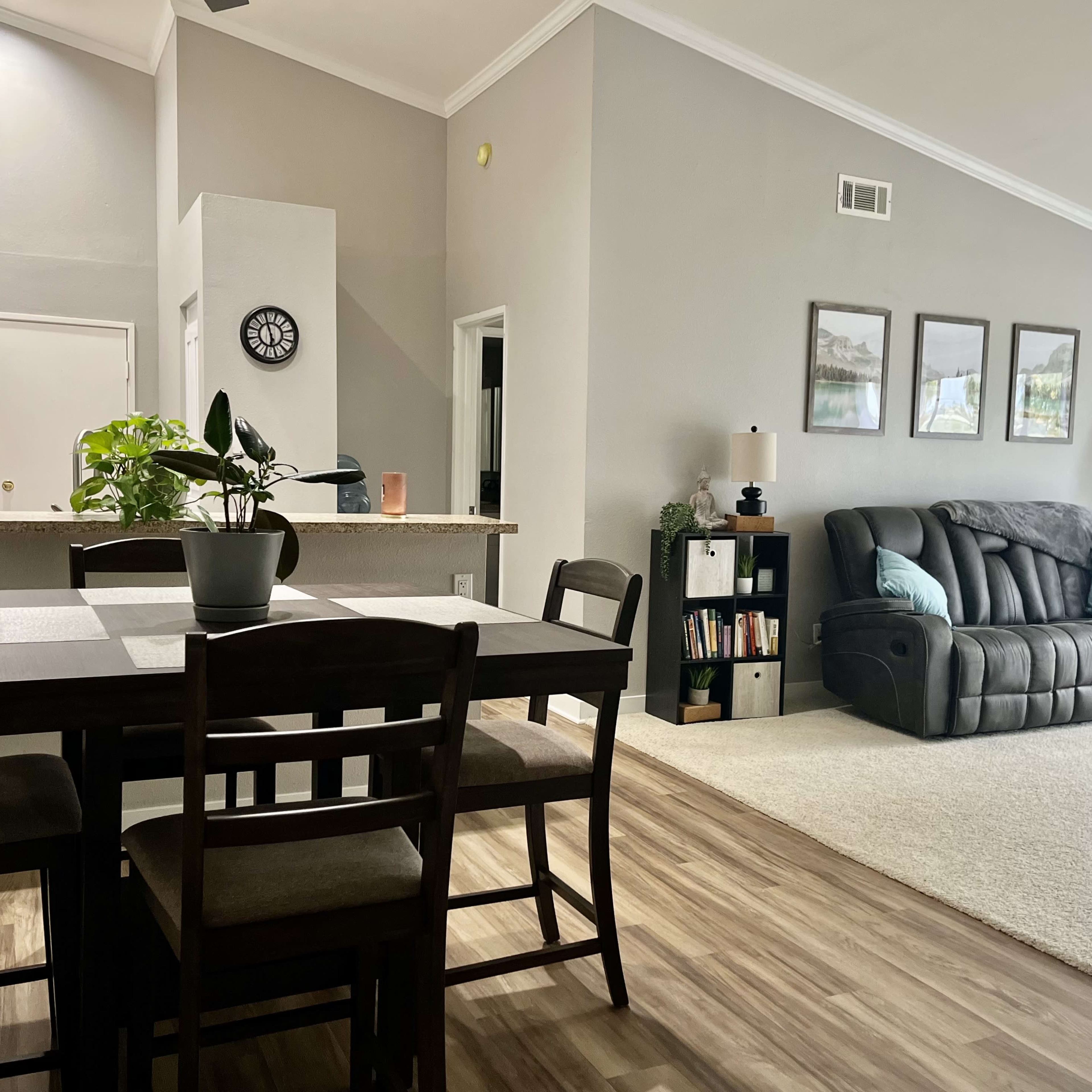 A spacious living area with a dining table, a gray sofa, and a large window, featuring neutral-colored walls and wood flooring.