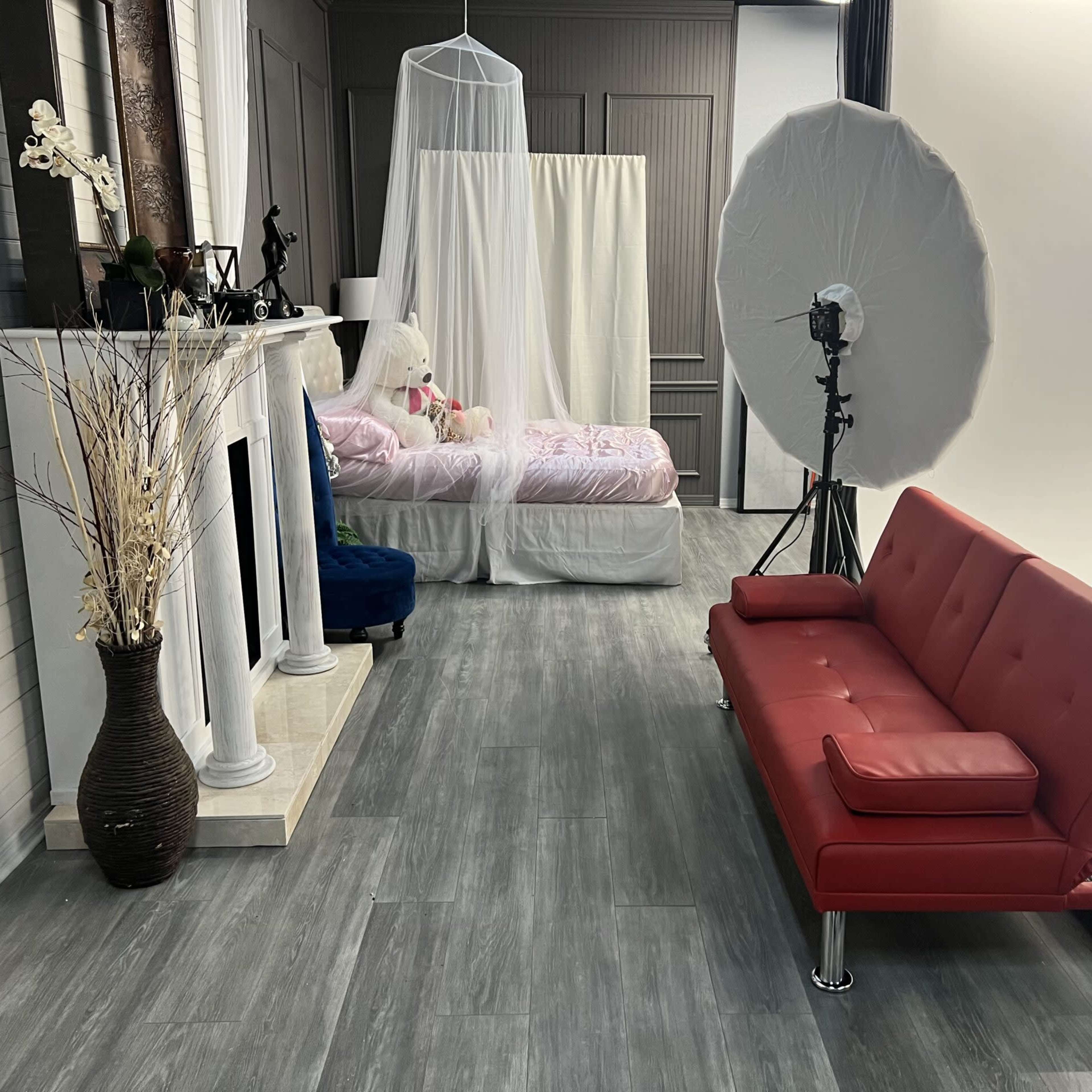 A studio room with a red sofa, a bed draped in white fabric, and a large lighting umbrella setup beside a decorative vase.
