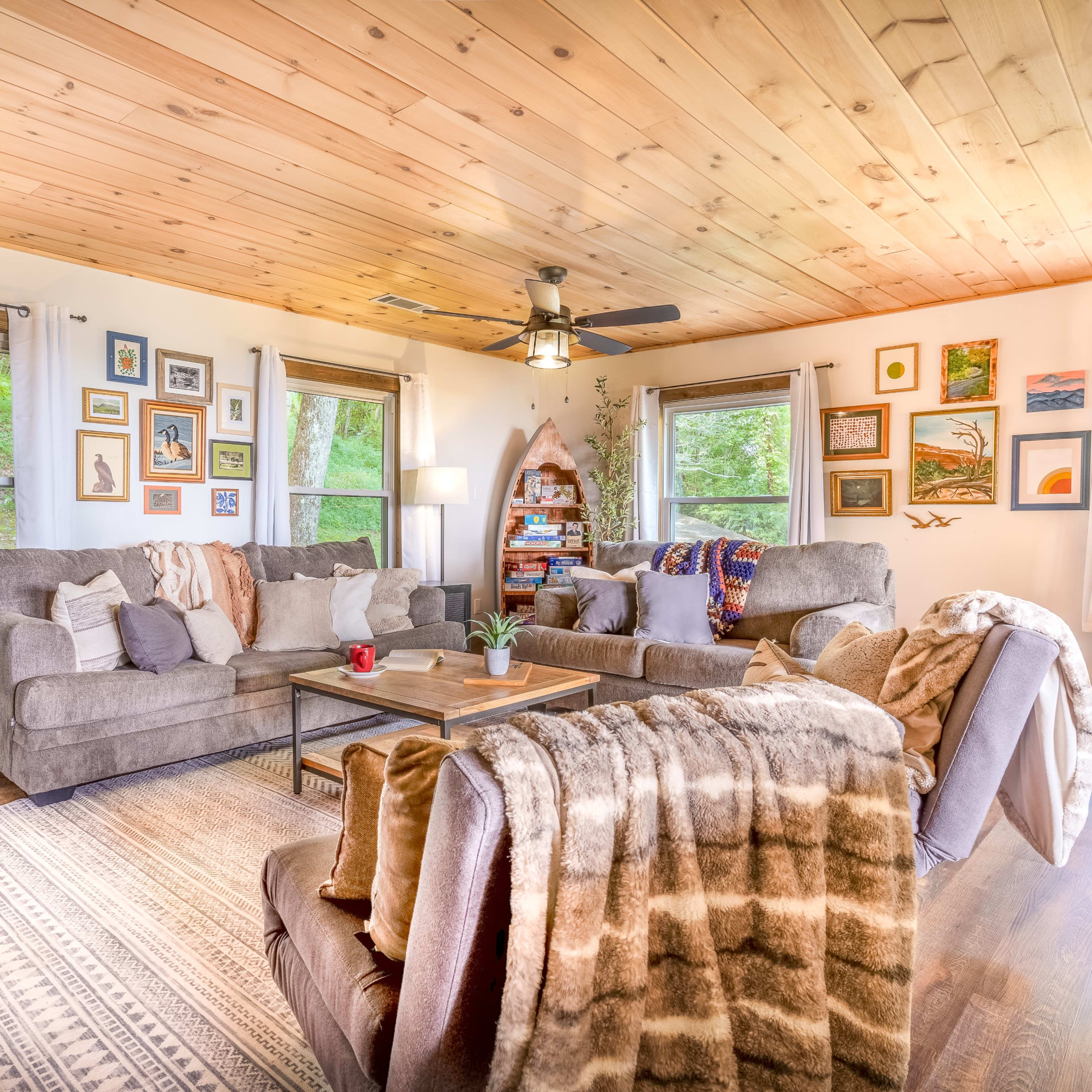 A cozy living room with a wooden ceiling, two large sofas, a coffee table, and various framed pictures on the walls, surrounded by windows letting in natural light.