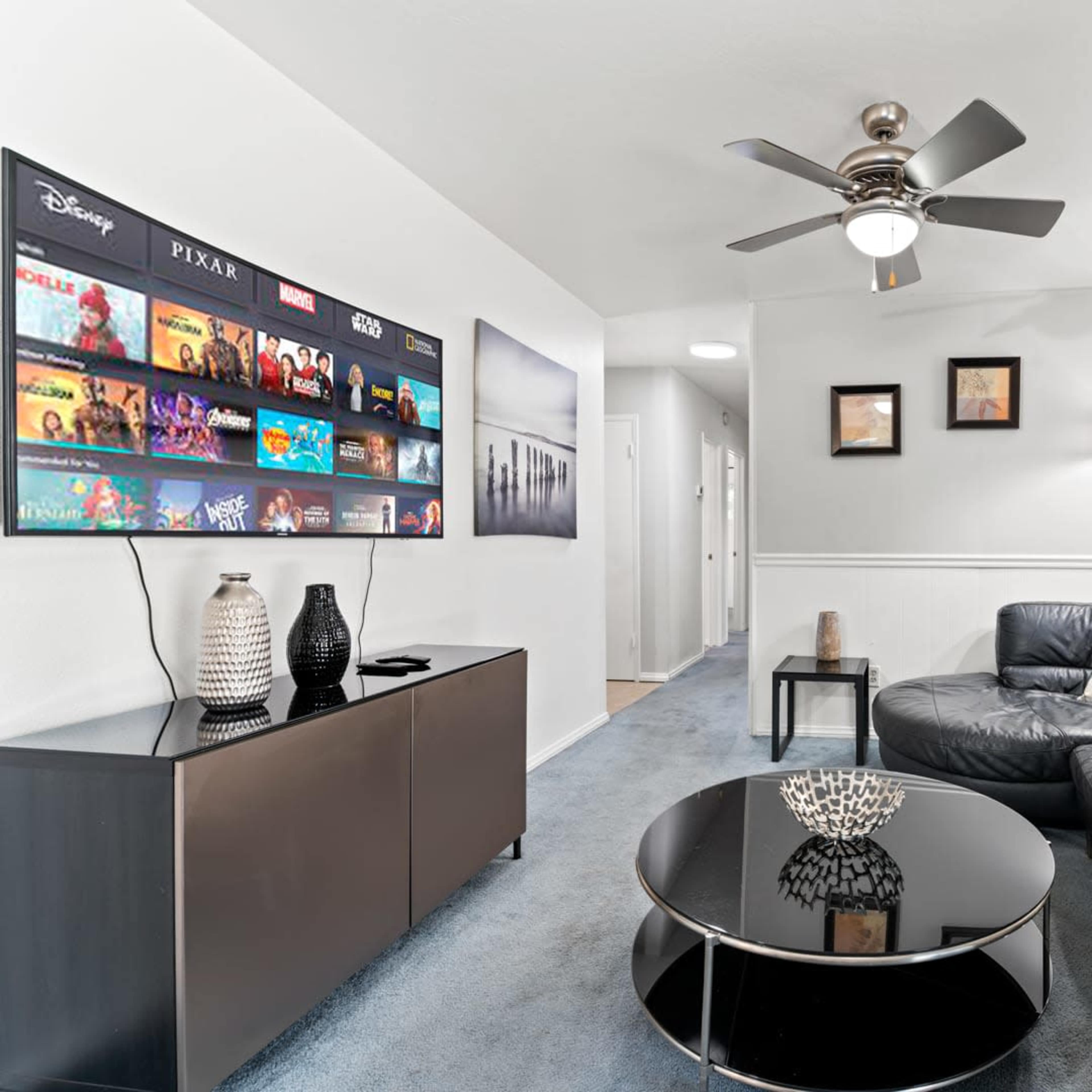 A modern living room features a black leather sectional couch, a round coffee table, and a wall-mounted TV displaying various movie titles.