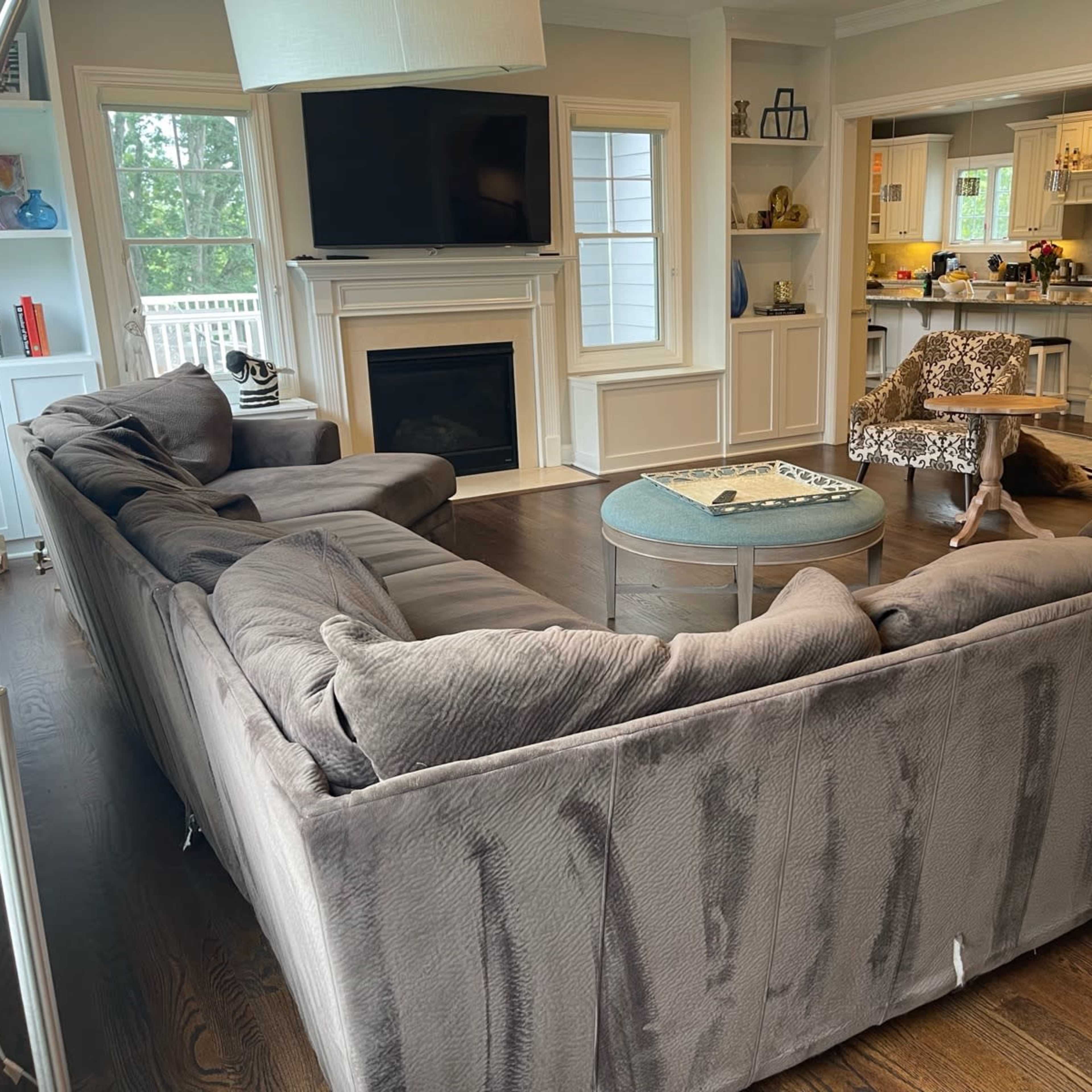A spacious living room with a large gray sectional sofa, two patterned chairs, a round coffee table, a fireplace, and a kitchen area visible in the background.