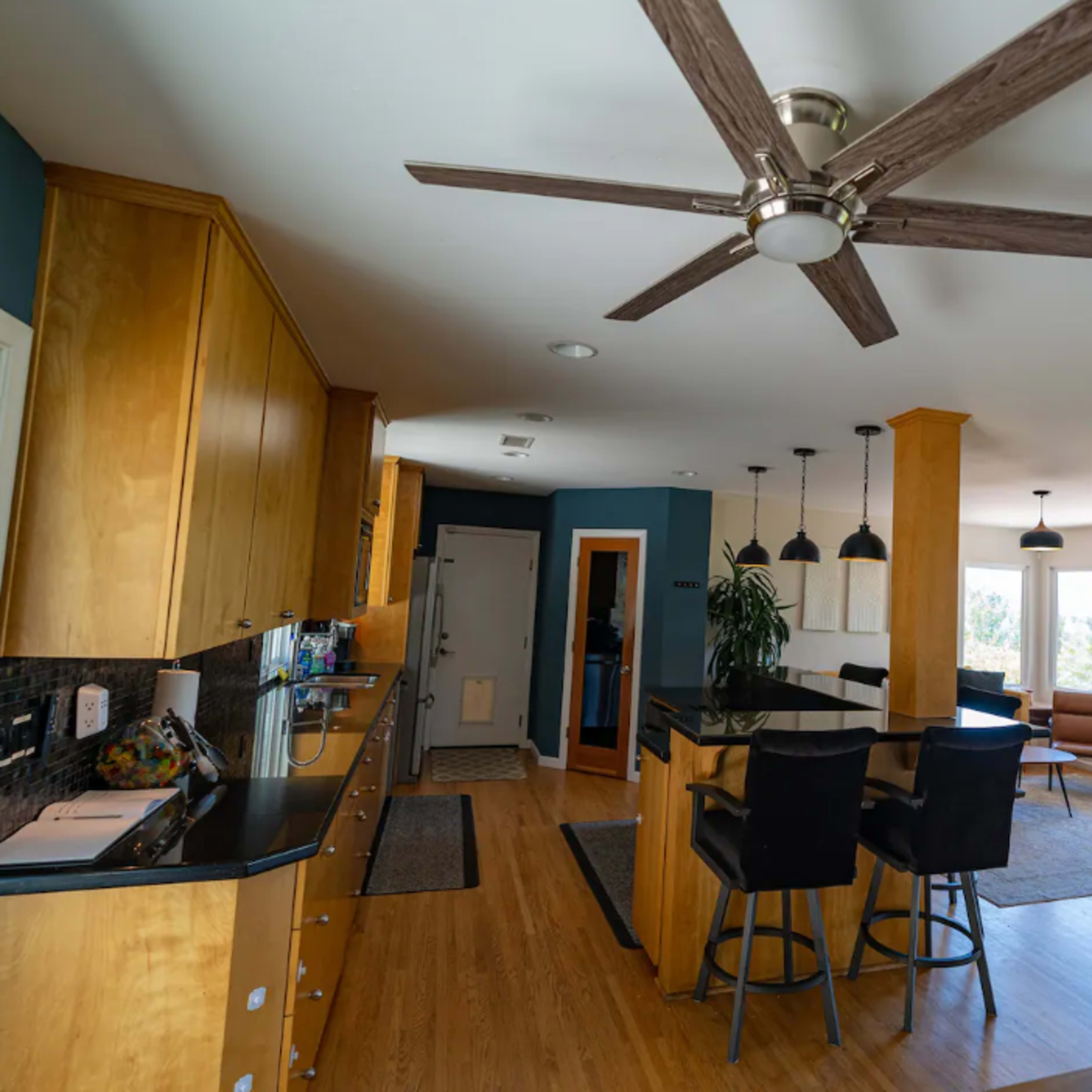 A modern kitchen and living area with wooden cabinets, a large island with bar stools, and a ceiling fan, featuring windows that provide a view of the outdoors.