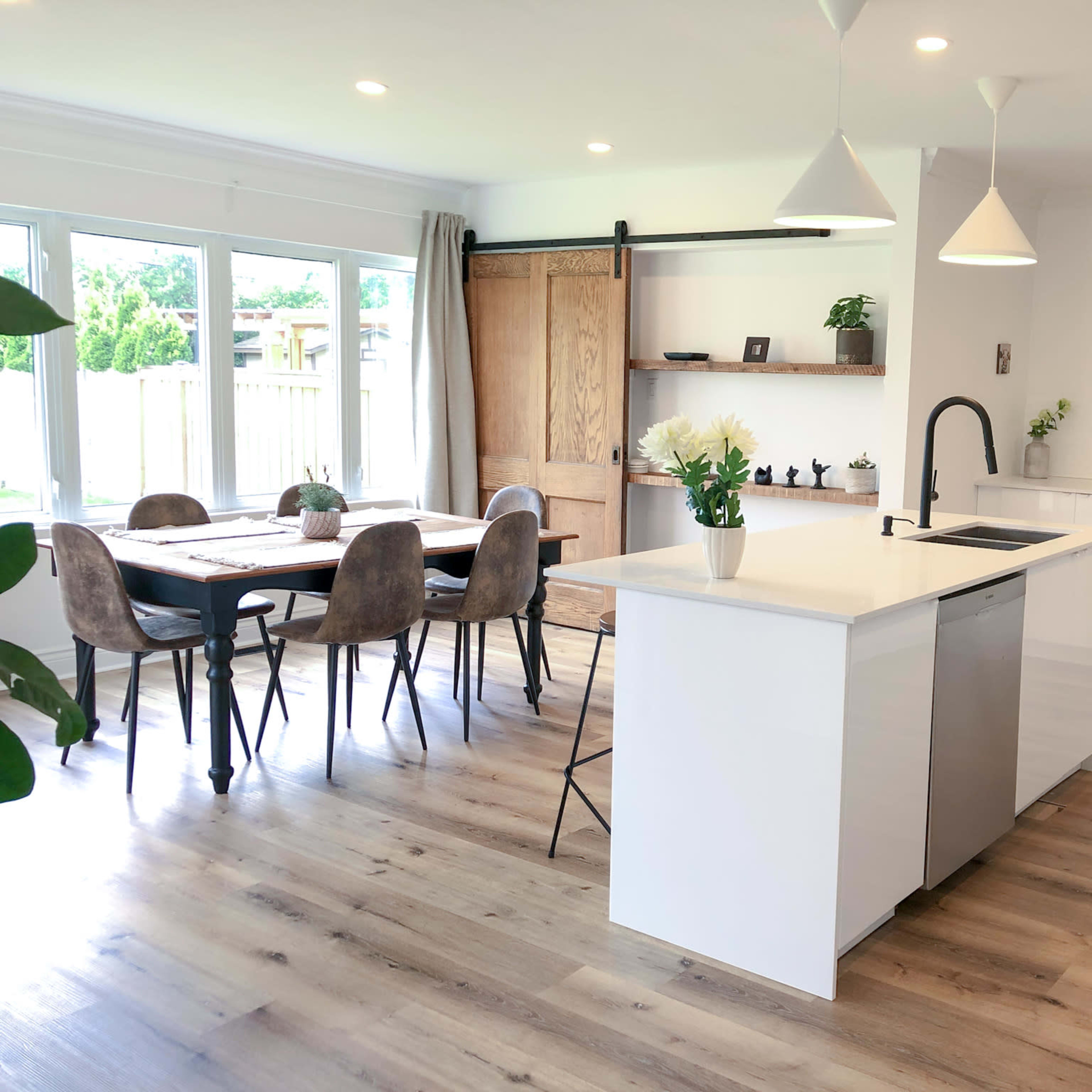 A modern kitchen features a central island with seating, stainless steel appliances, a wooden dining table, and large windows allowing natural light.