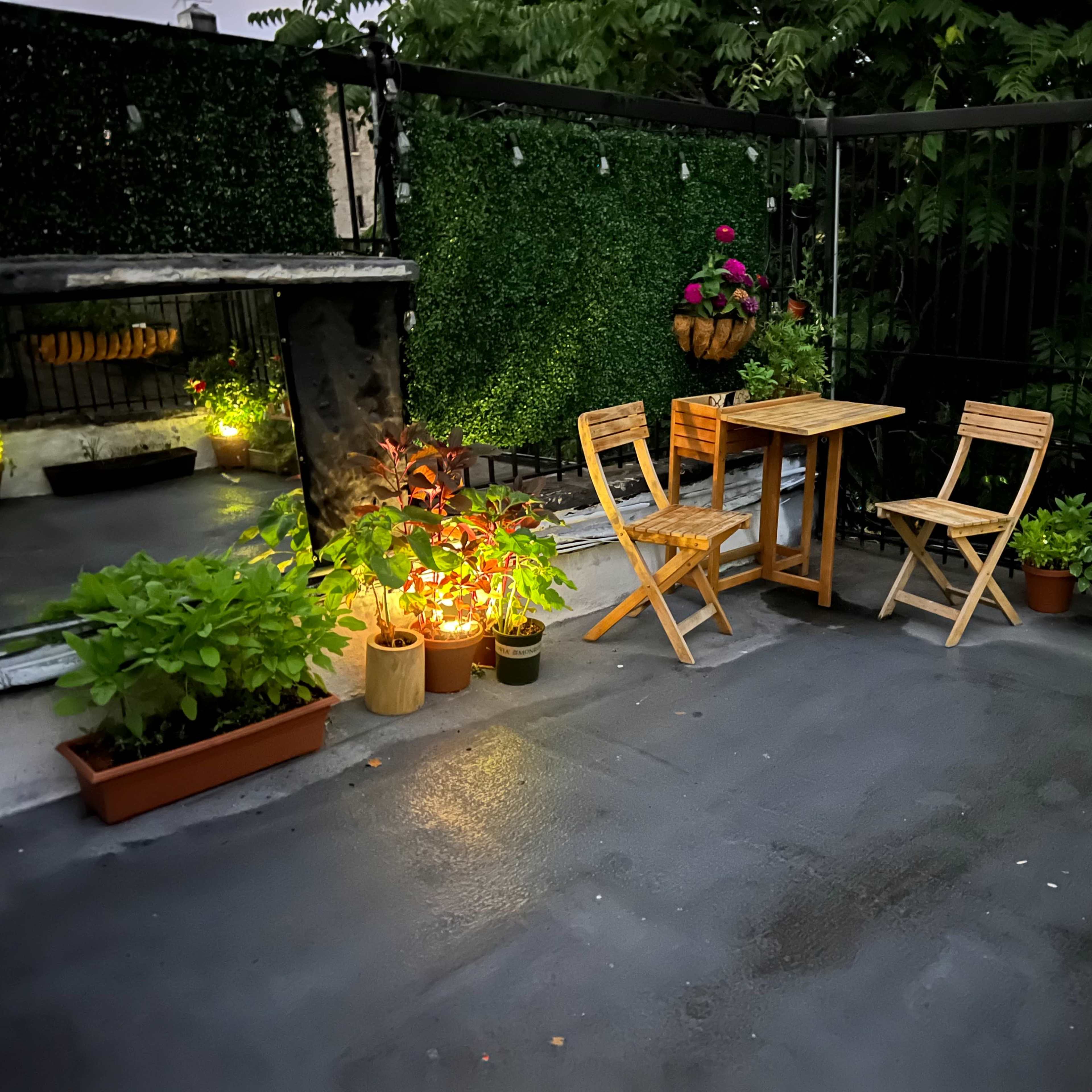 A rooftop terrace with two wooden chairs and a small table surrounded by potted plants and soft lighting.