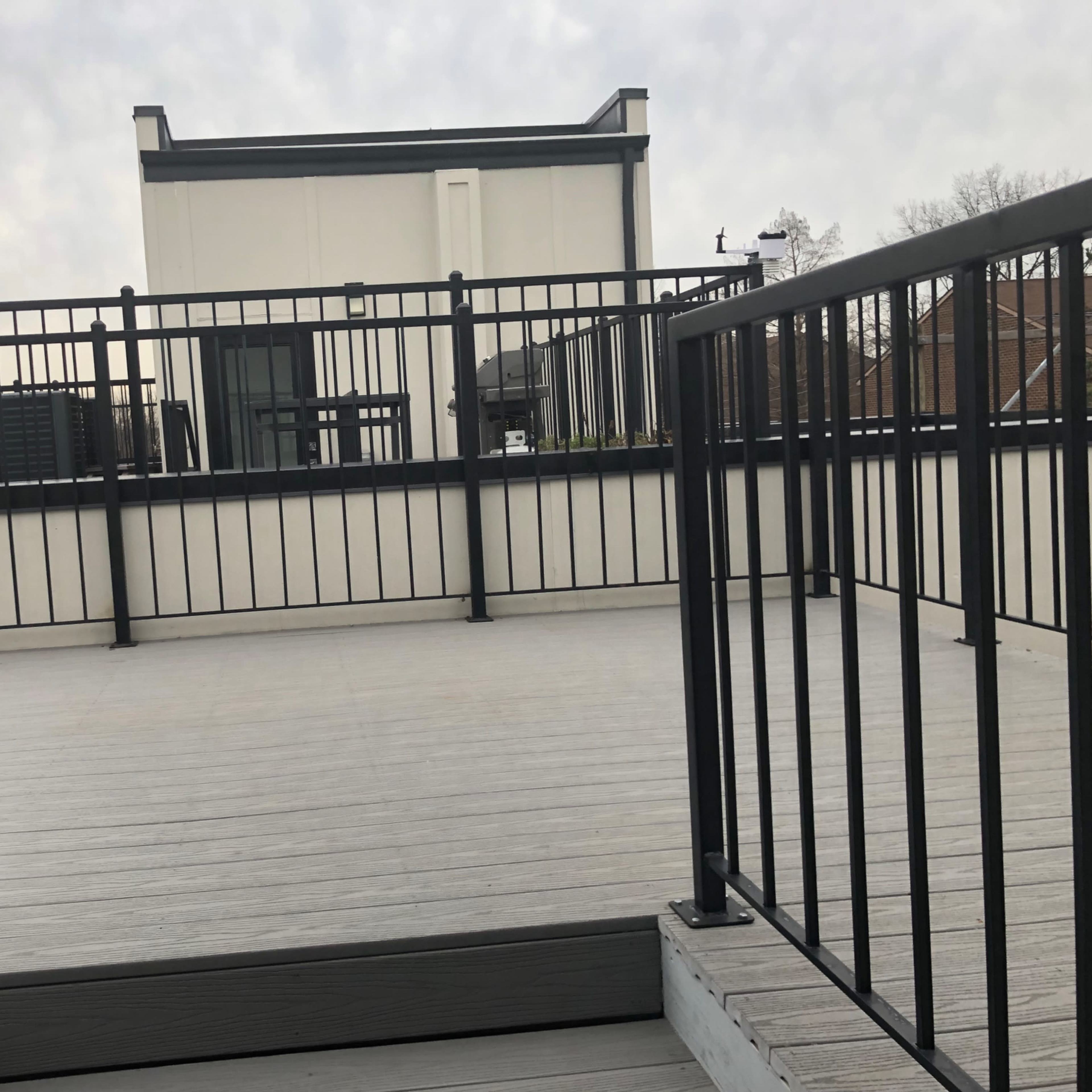 A rooftop deck with a wooden floor and black metal railings, leading towards a white building in the background.