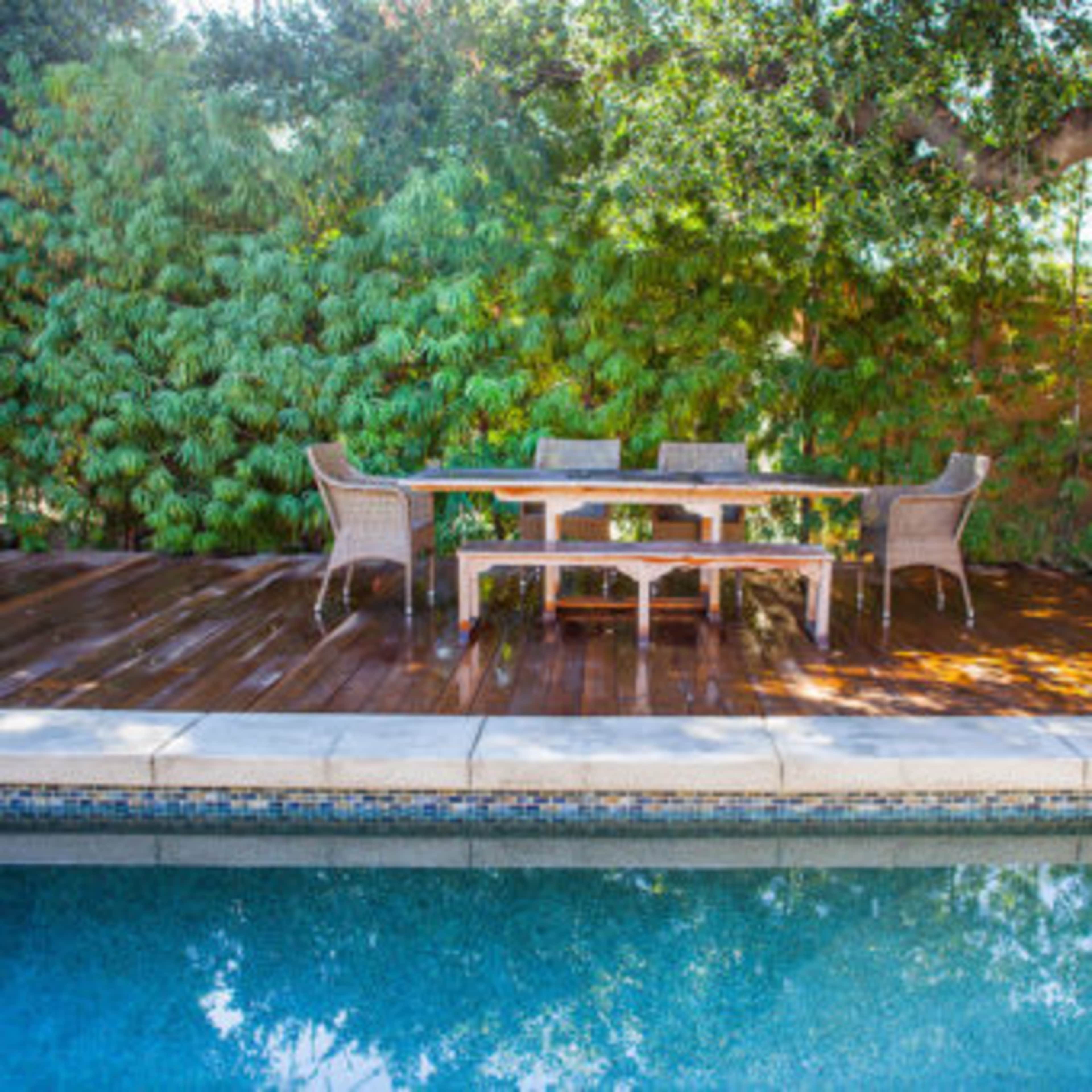A wooden deck with a dining table and chairs is positioned next to a swimming pool, surrounded by lush greenery.