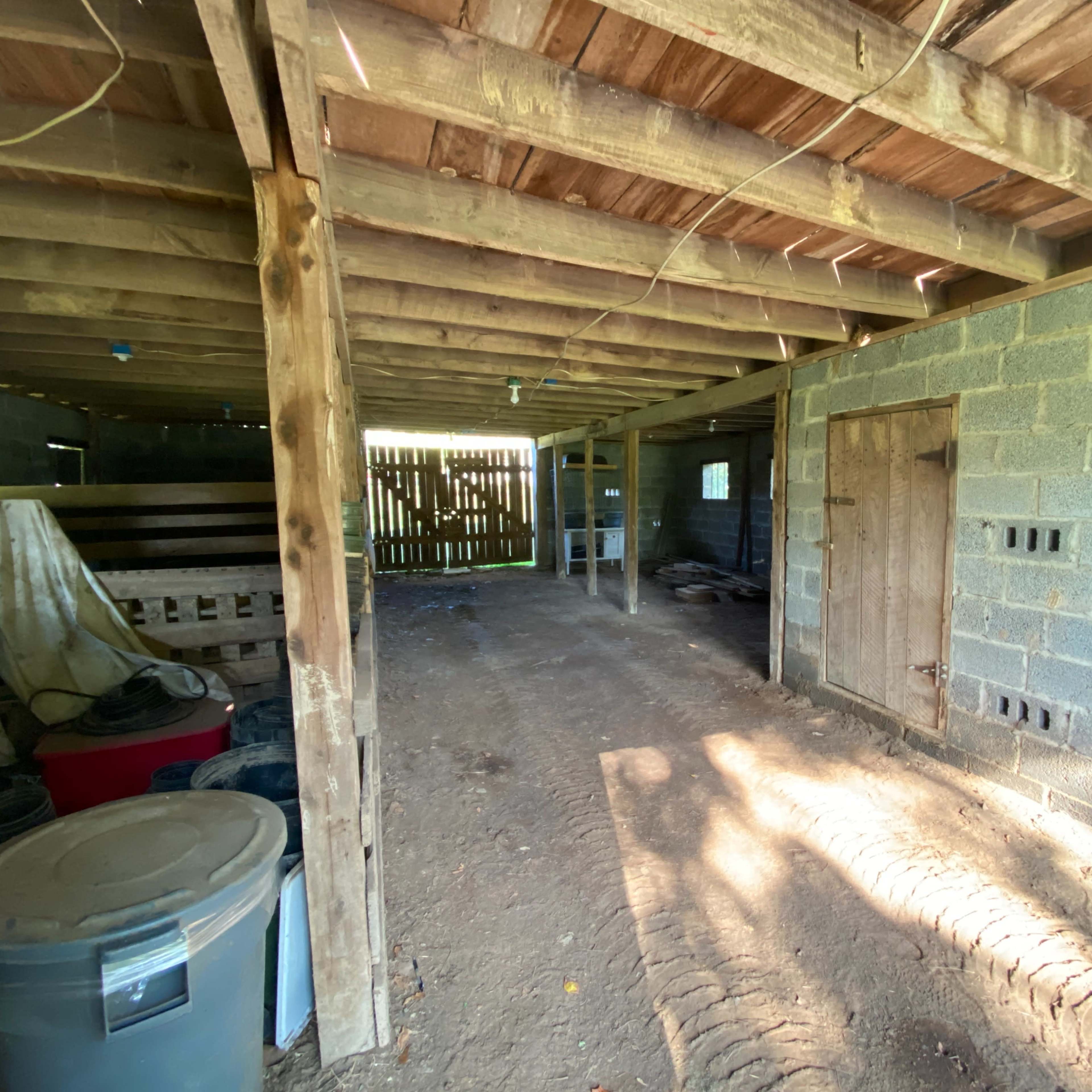 The interior of a rustic barn with exposed wooden beams, concrete walls, and dirt flooring, featuring an empty space on one side and a wooden door on the other.