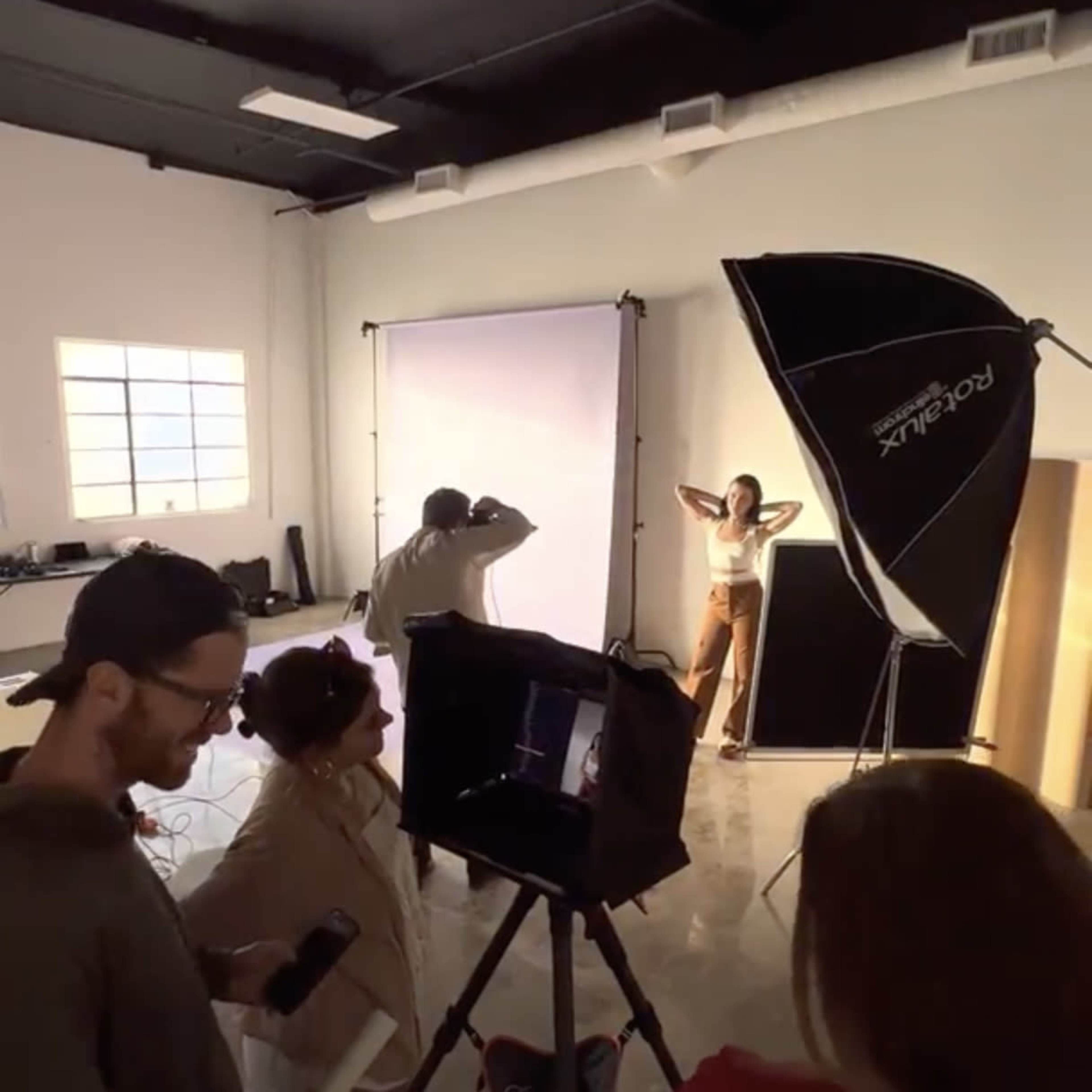 A group of people works in a photography studio, with a model posing under bright lights while photographers and assistants adjust equipment.