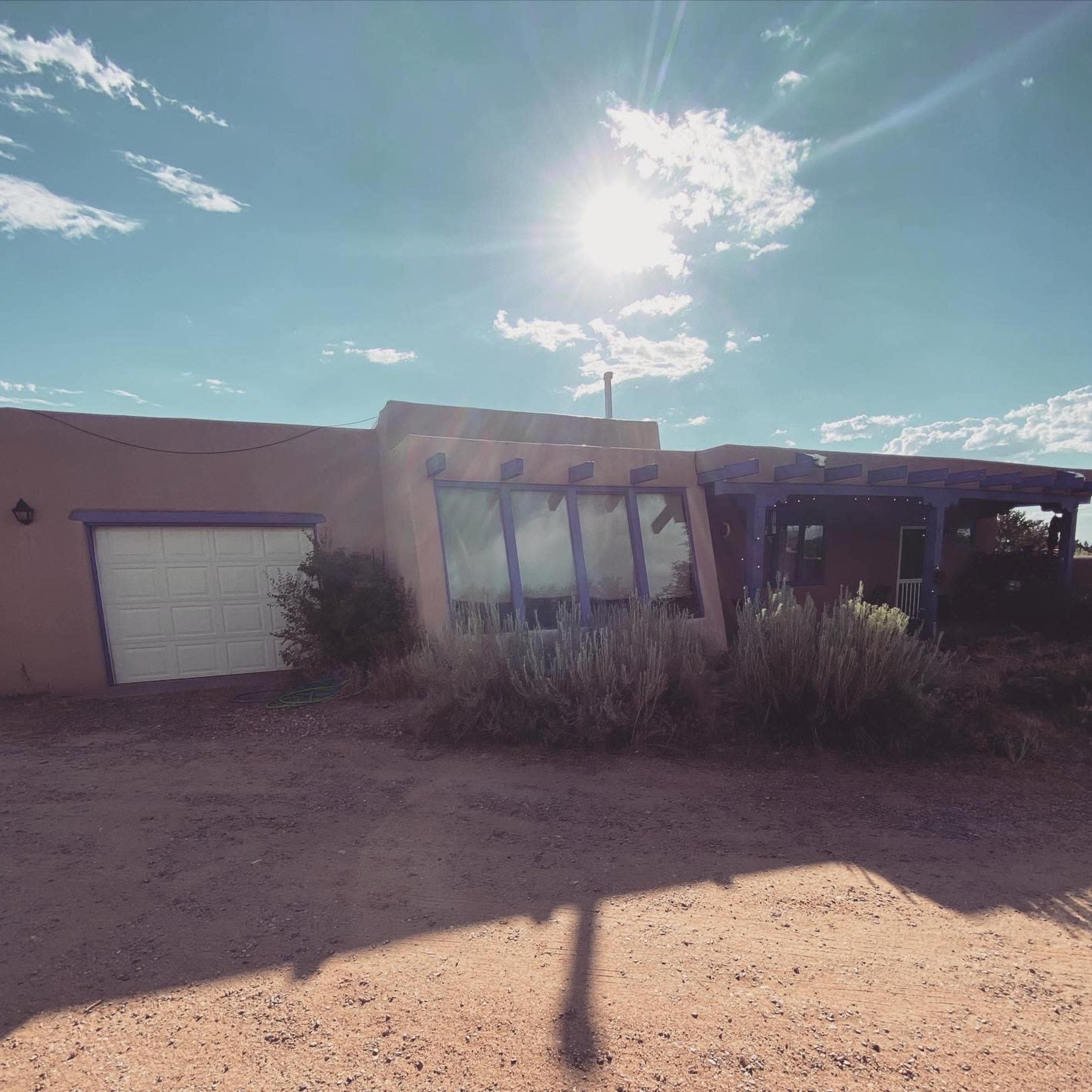 A single-story adobe house with large windows and a garage door is set against a clear sky with the sun shining above.