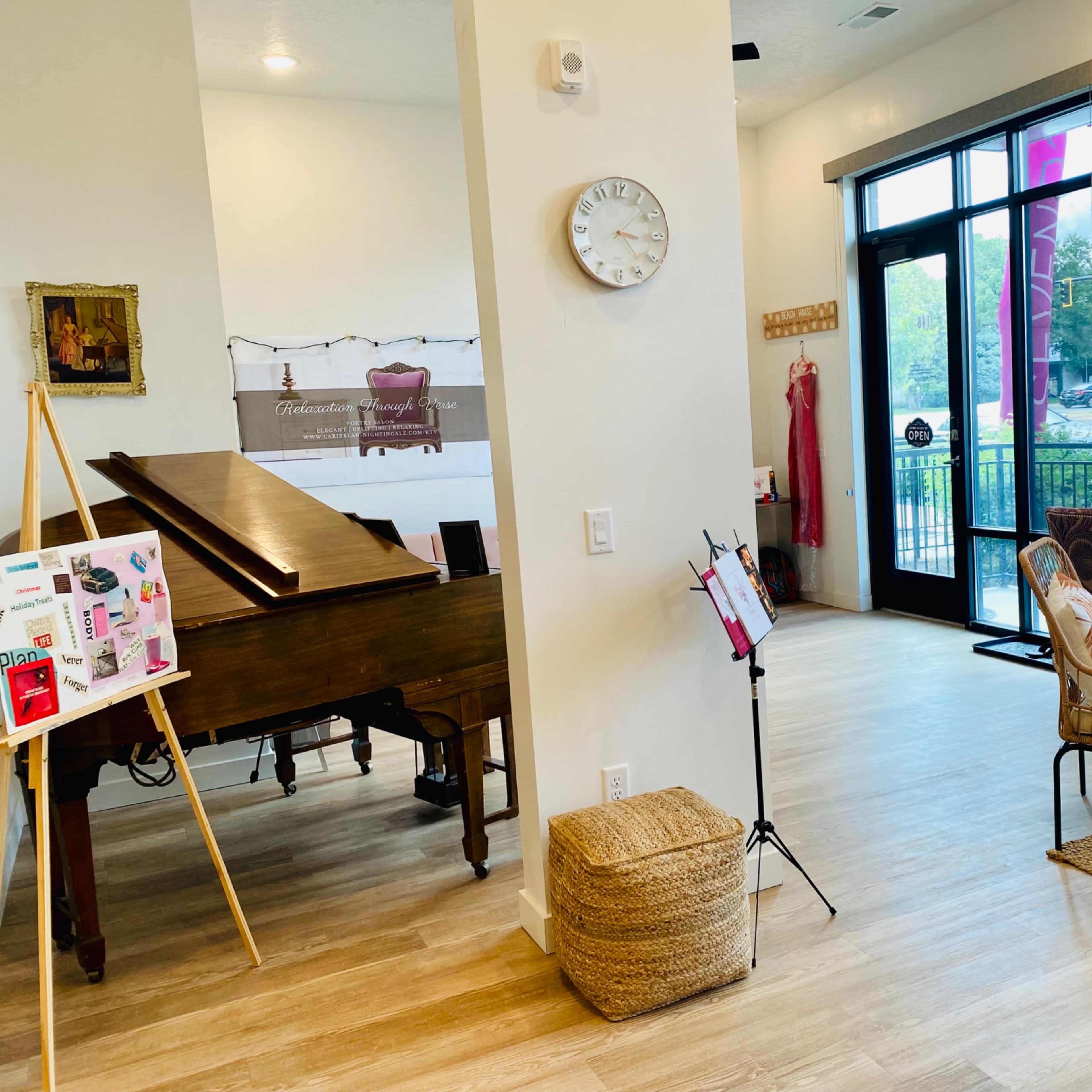 An indoor space featuring a grand piano, a wall clock, a wicker chair, and large windows with a view outside.