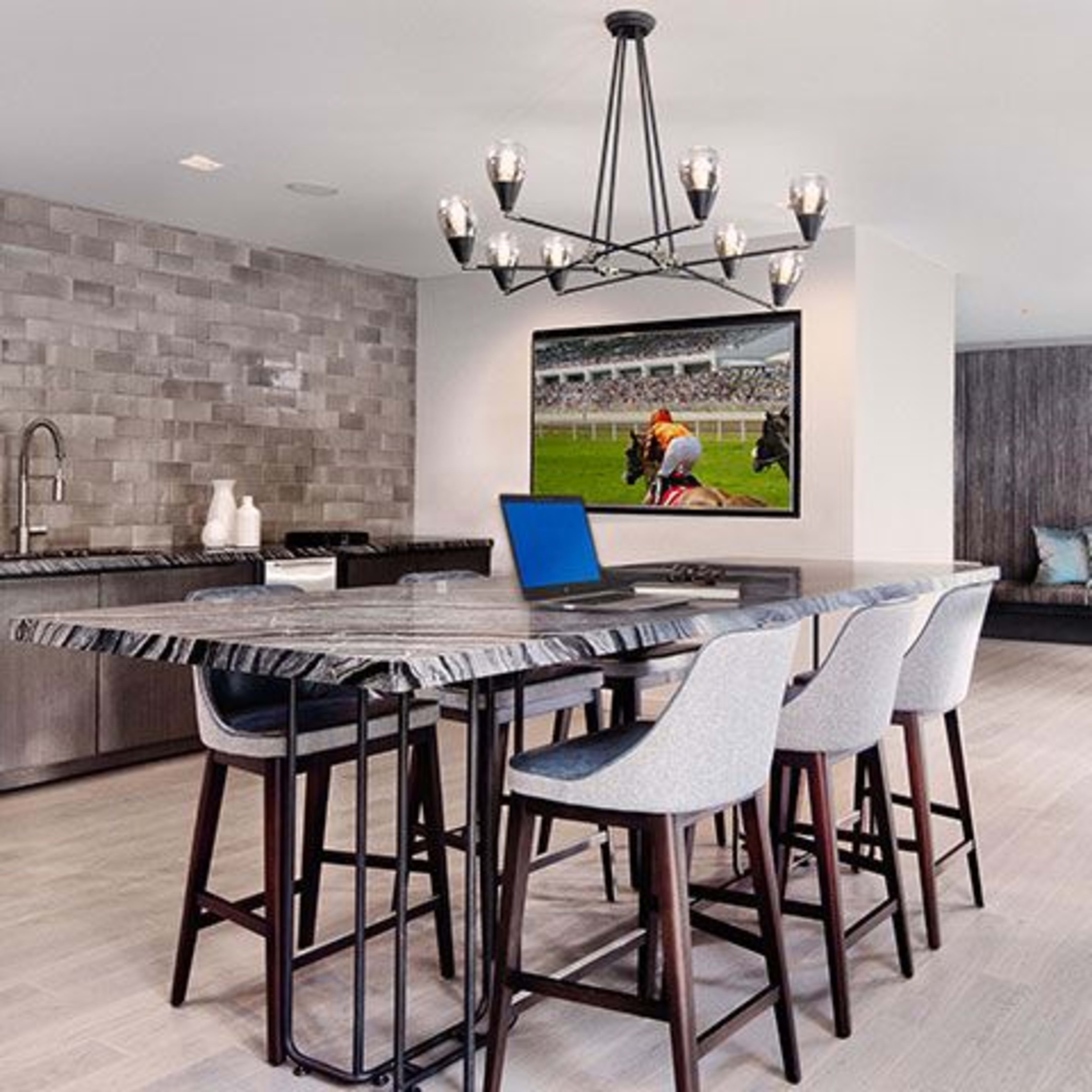 A modern kitchen or dining area with a large island, several high stools, a wall-mounted television displaying a sports event, and a cozy seating area in the background.