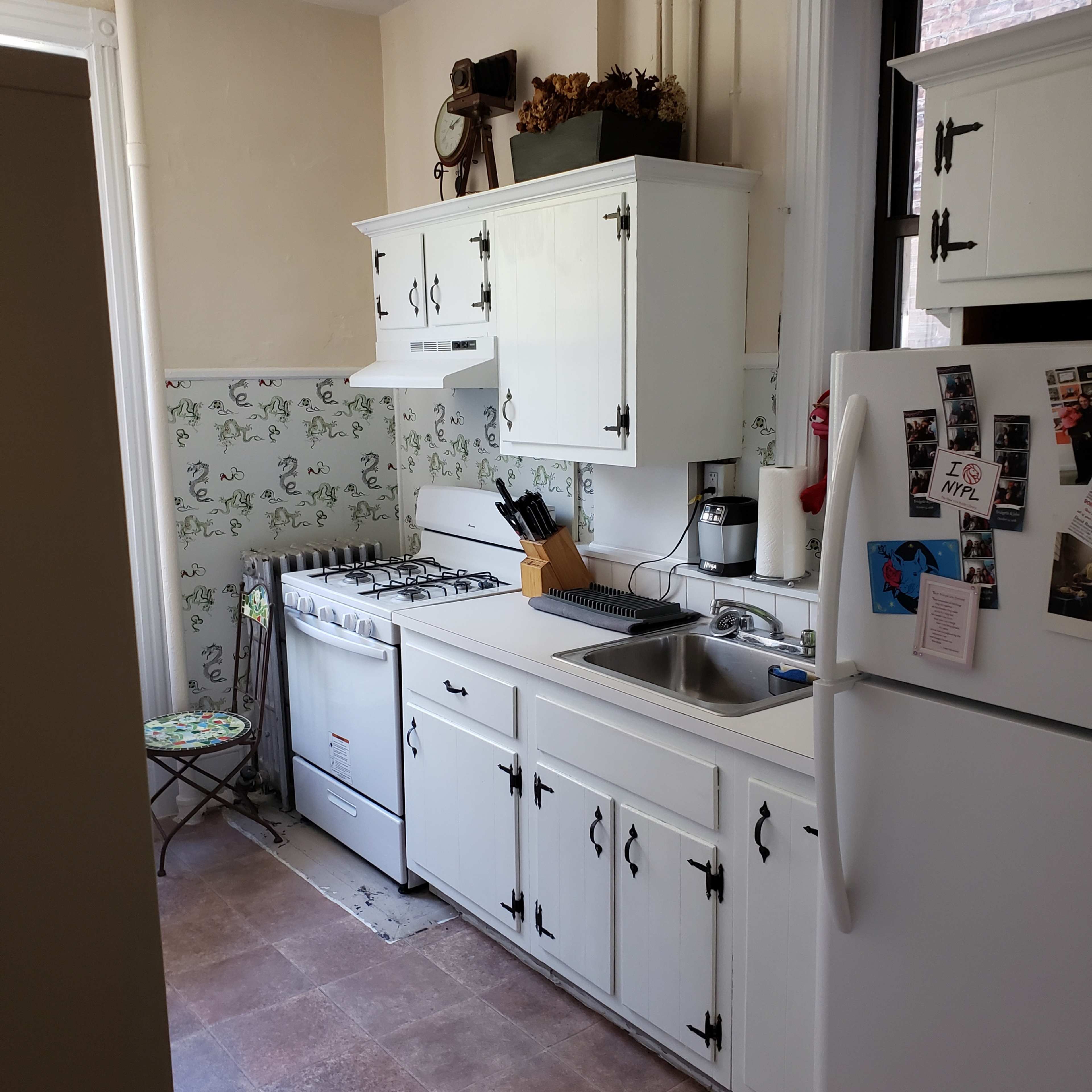 A small kitchen with white cabinets, a stove, sink, and refrigerator, along with patterned wallpaper and a tiled floor.