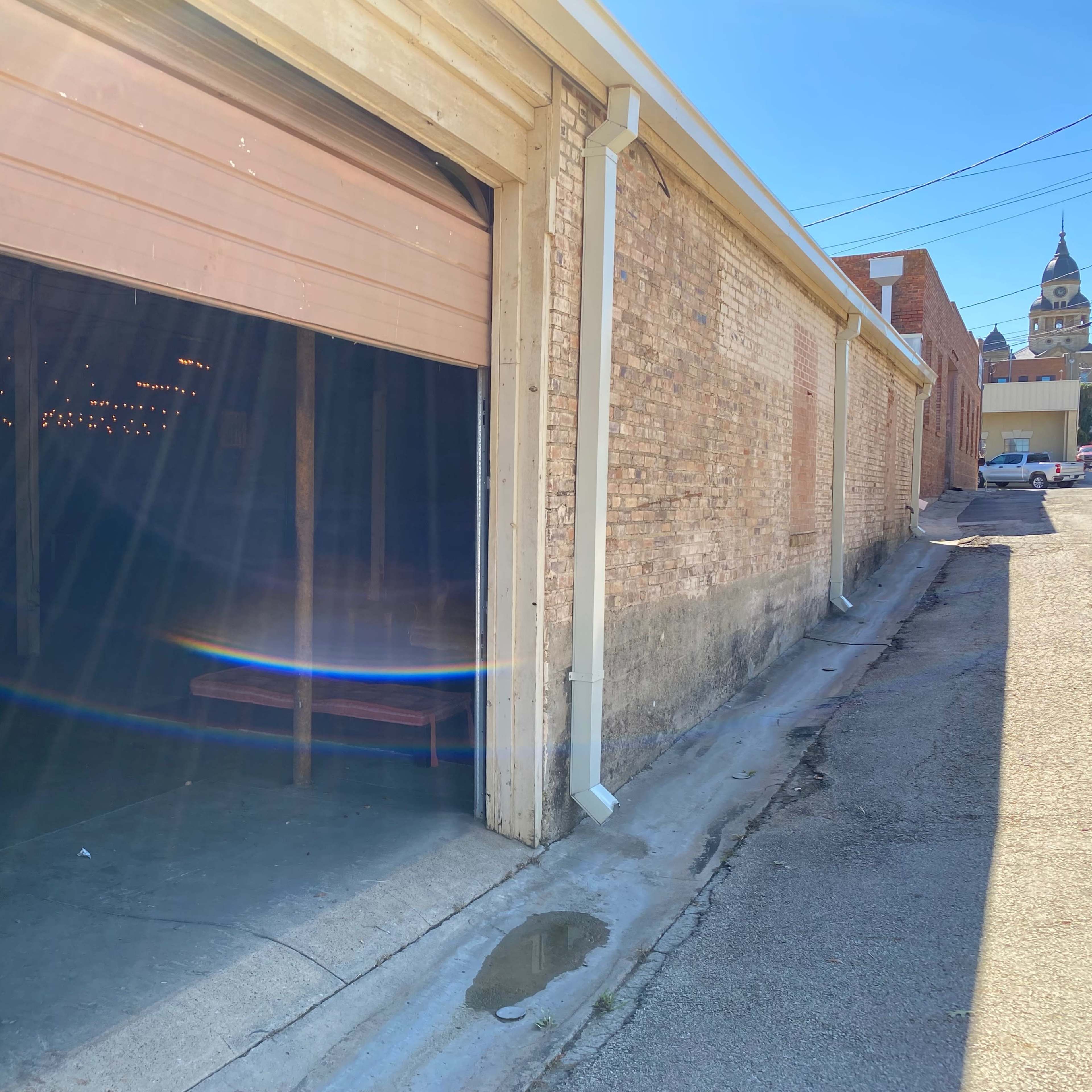 An open garage door reveals a dimly lit interior with string lights visible inside, set against a brick wall in a narrow alleyway.