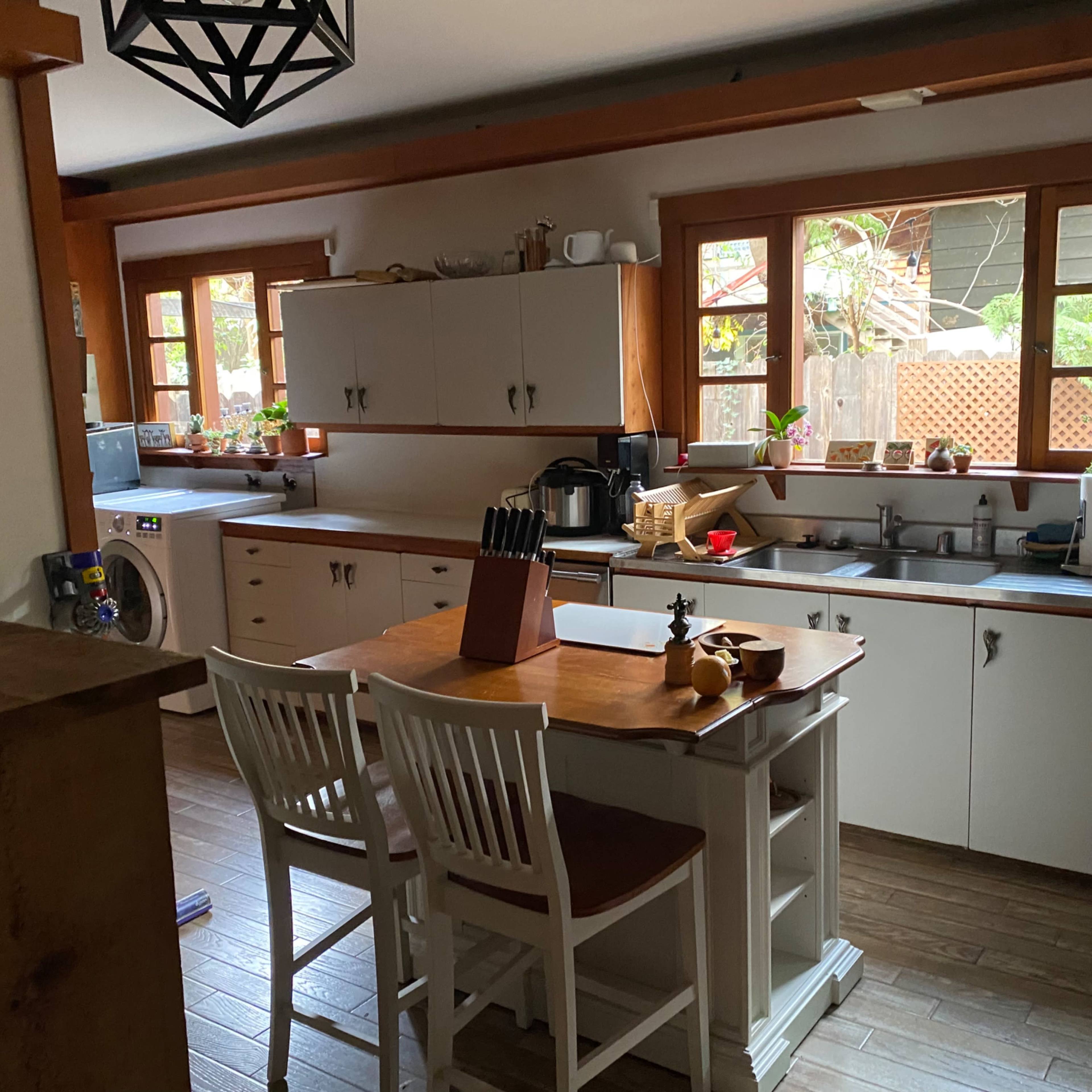 A kitchen with white cabinets, a wooden table with chairs, and windows allowing natural light to enter.