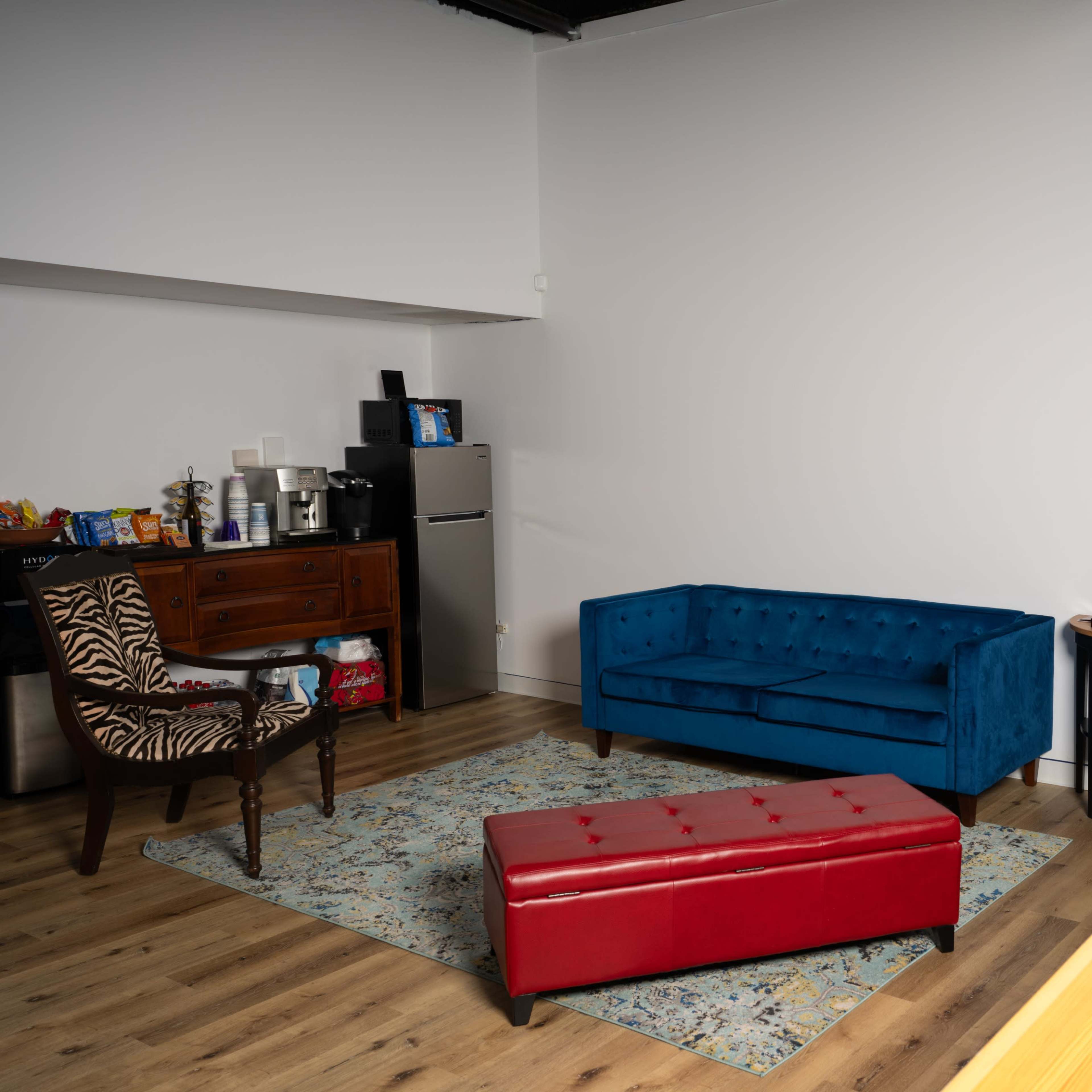 A modern room with a blue tufted sofa, a red ottoman, a wooden chair with a patterned upholstery, and a small table, all set against a backdrop of a brick wall.