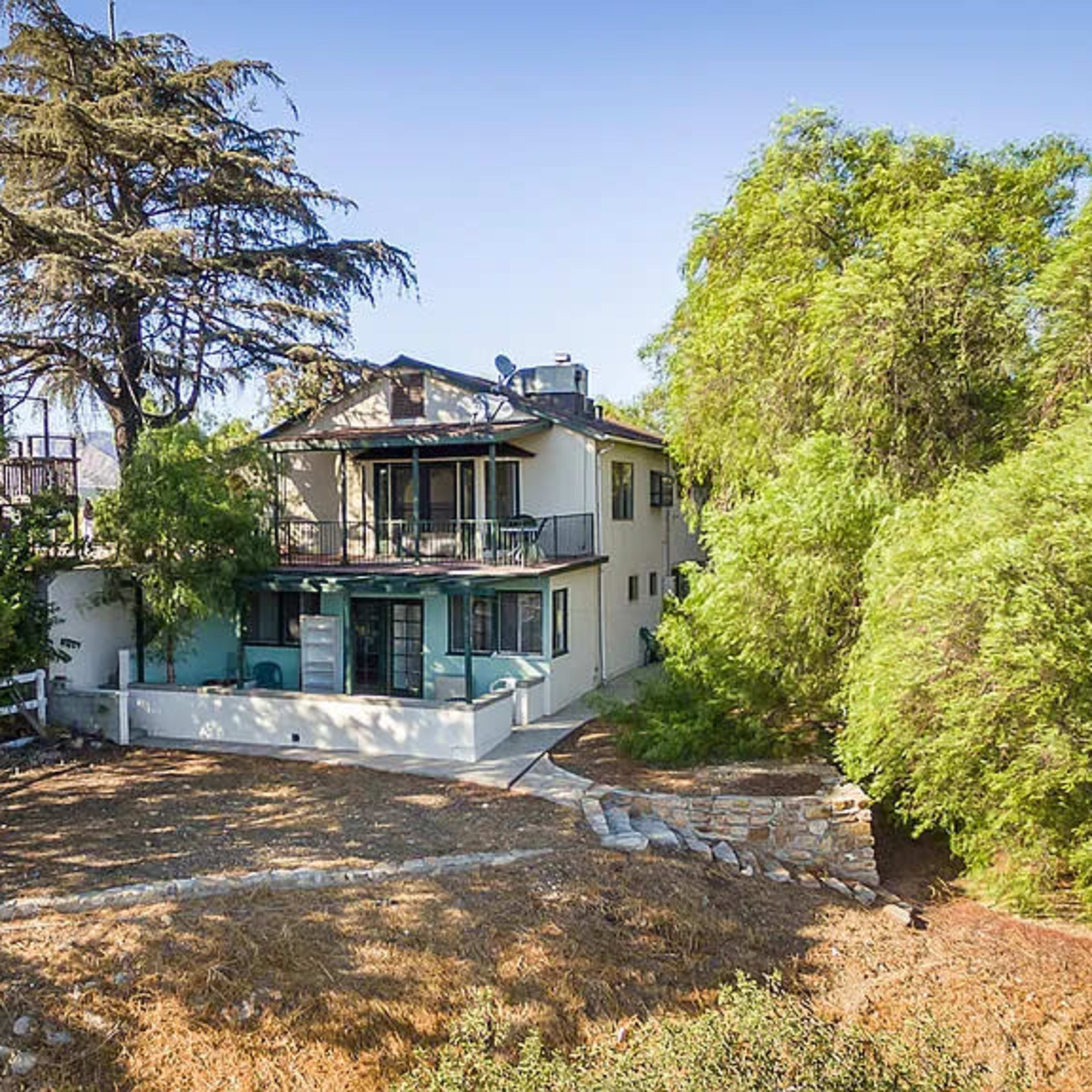 A two-story house surrounded by trees, with a patio and a stone pathway leading to it.