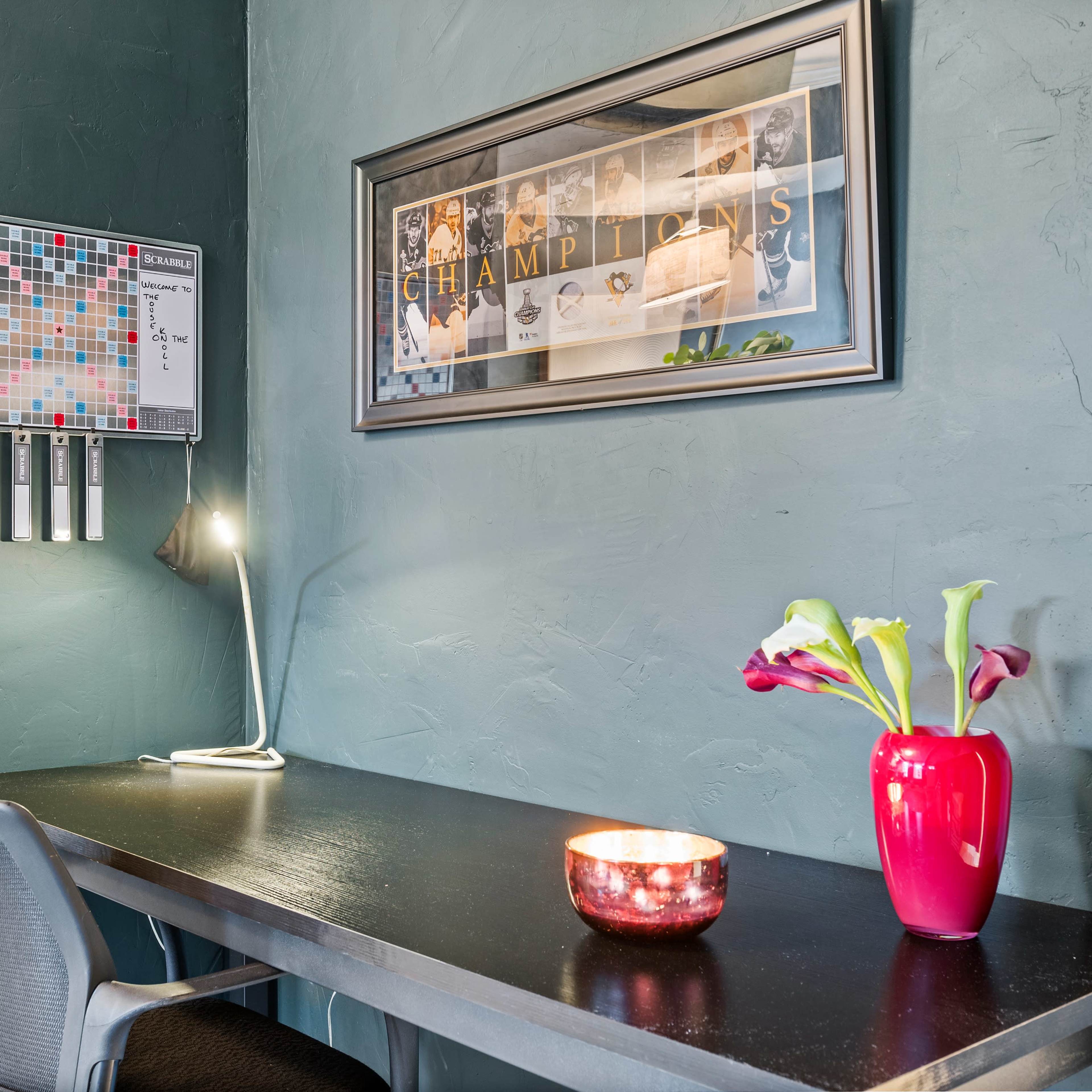A small desk with a lamp, a framed picture on the wall, a decorative vase with flowers, and a candle on the surface against a textured green wall.