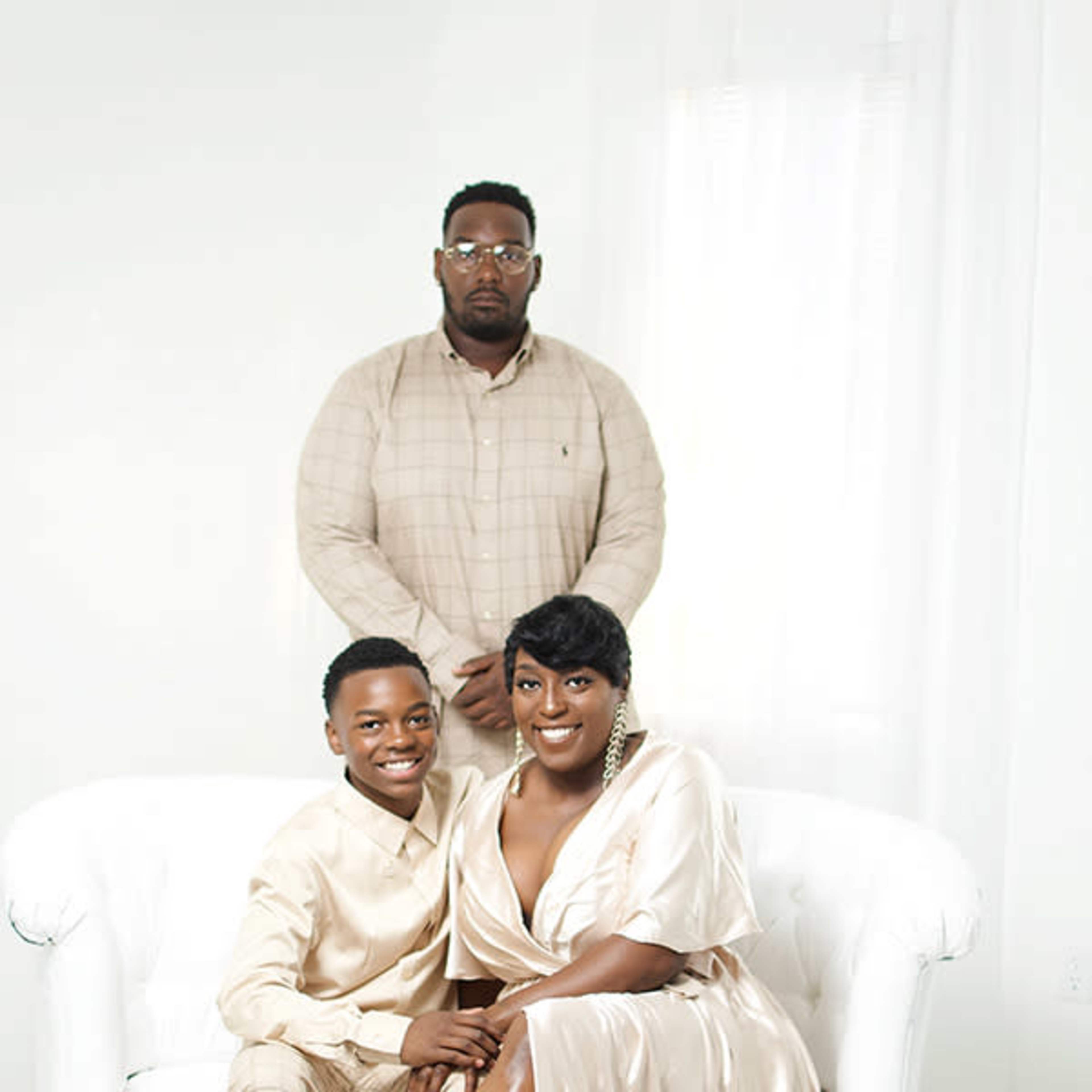 A man and woman, both dressed in formal attire, are seated on a white couch, while another man stands behind them.