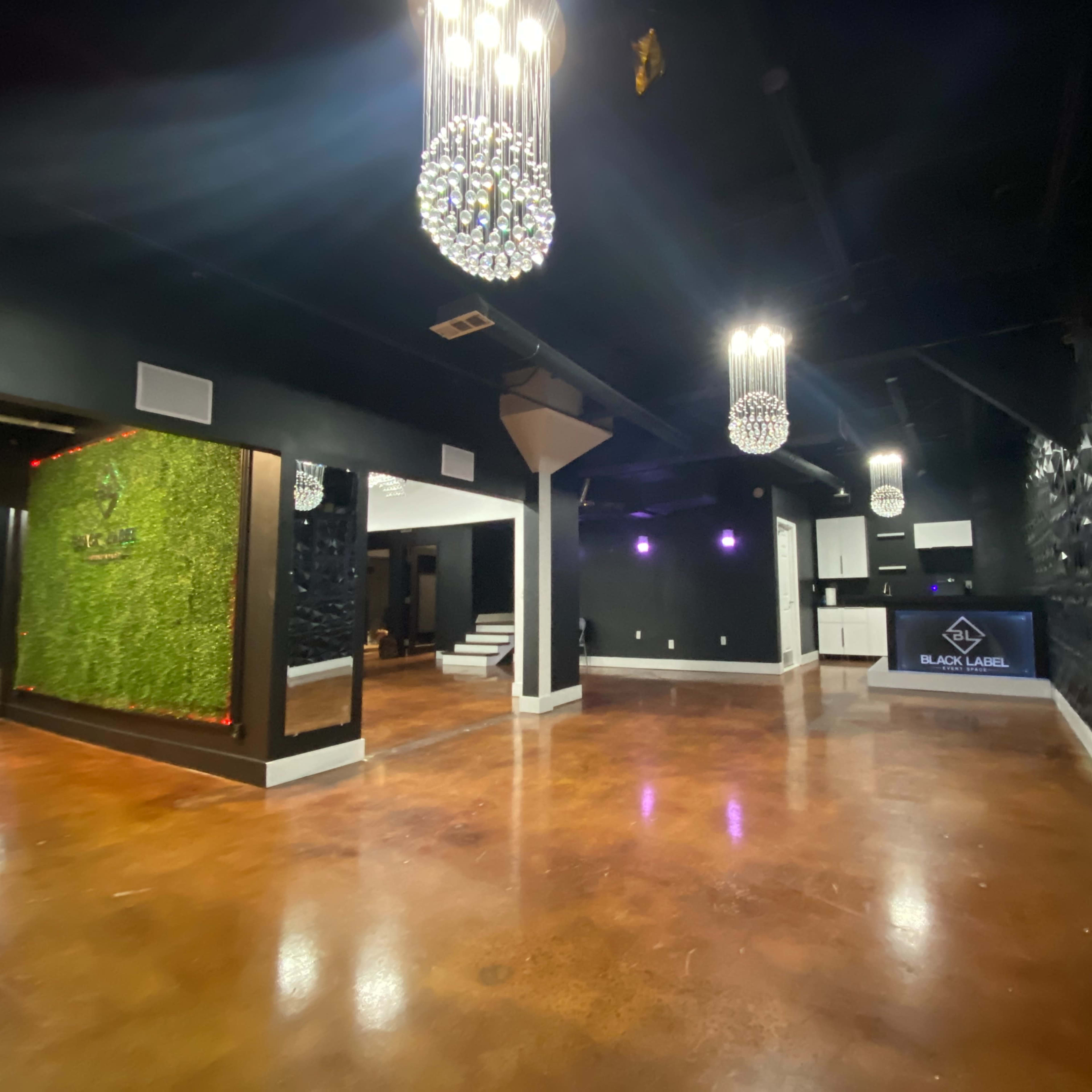 An empty, contemporary event space featuring black walls, decorative greenery, and pendant chandeliers.