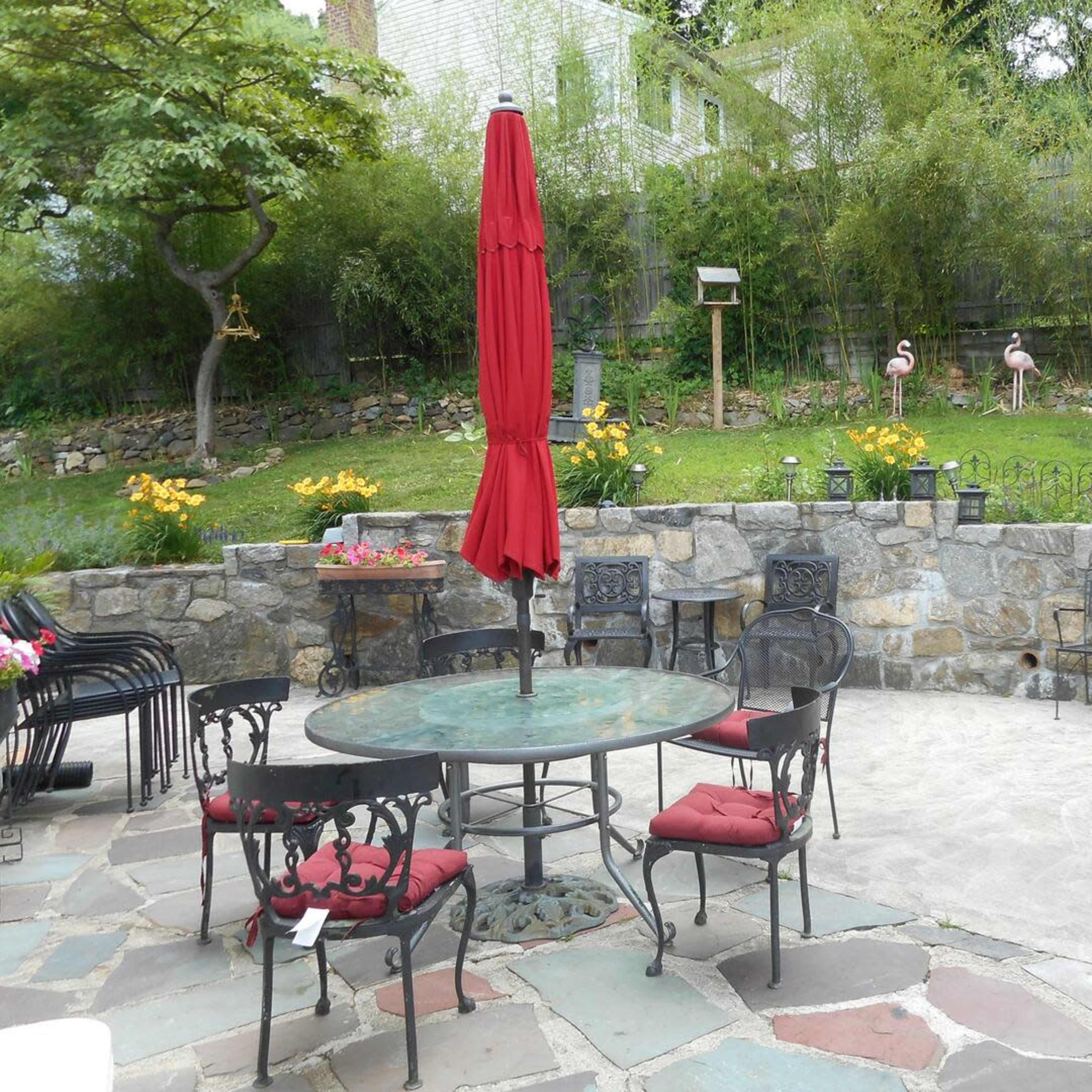 A stone patio with a round table and chairs, featuring a red umbrella, surrounded by a garden with flowers and decorative flamingos.