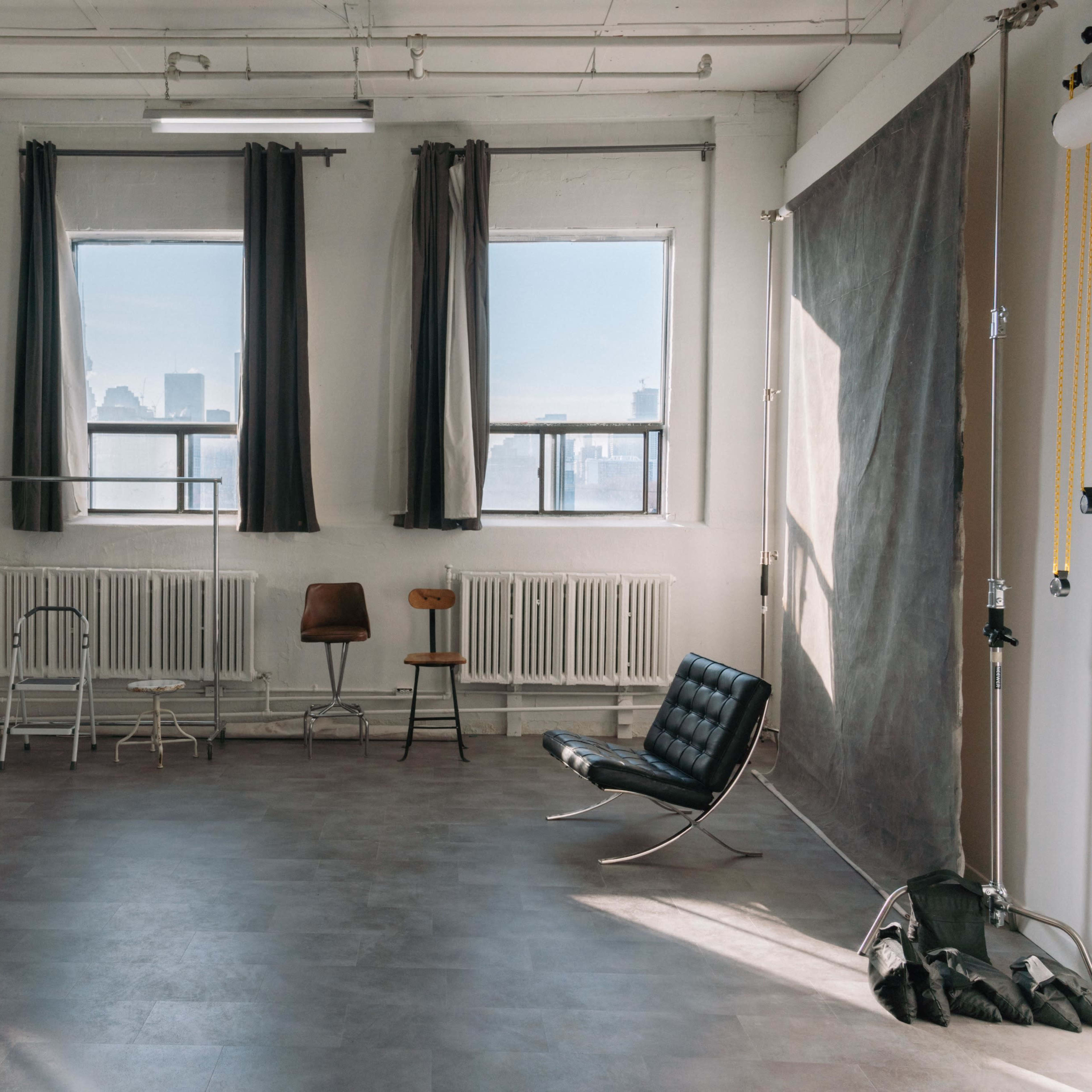 A spacious, sunlit studio features large windows, a minimalist black chair, and various pieces of furniture along the walls.