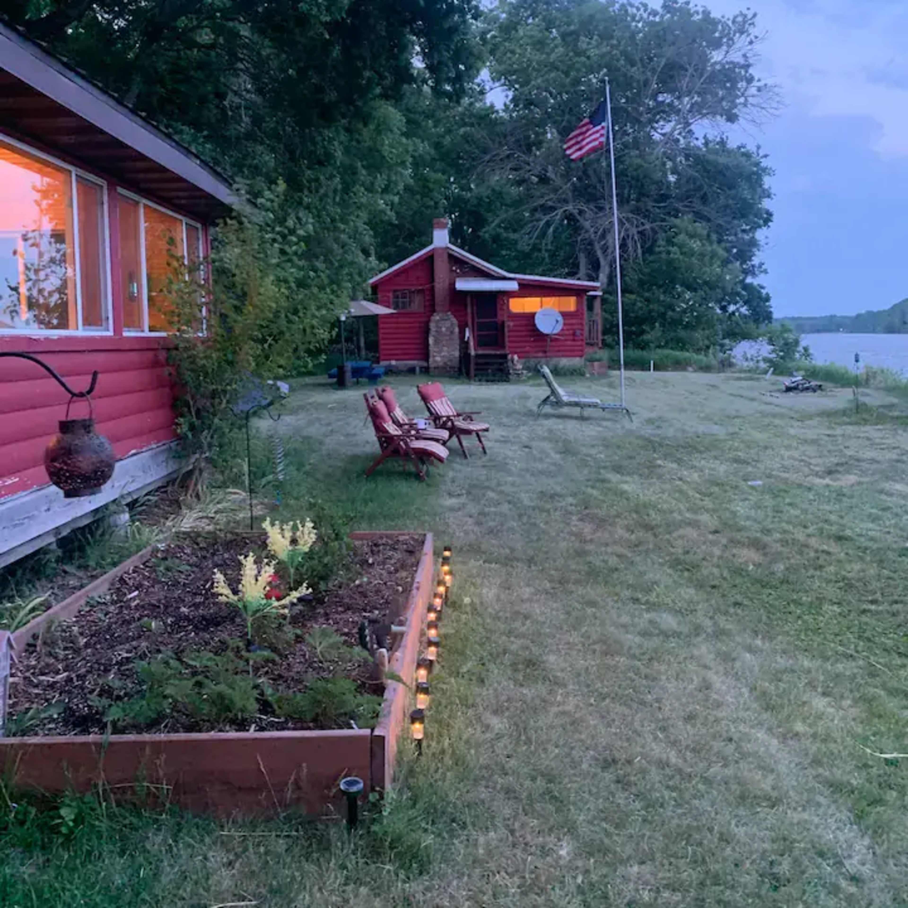 A red cabin with a flag stands near a river, accompanied by lounge chairs and a flower garden in the foreground.
