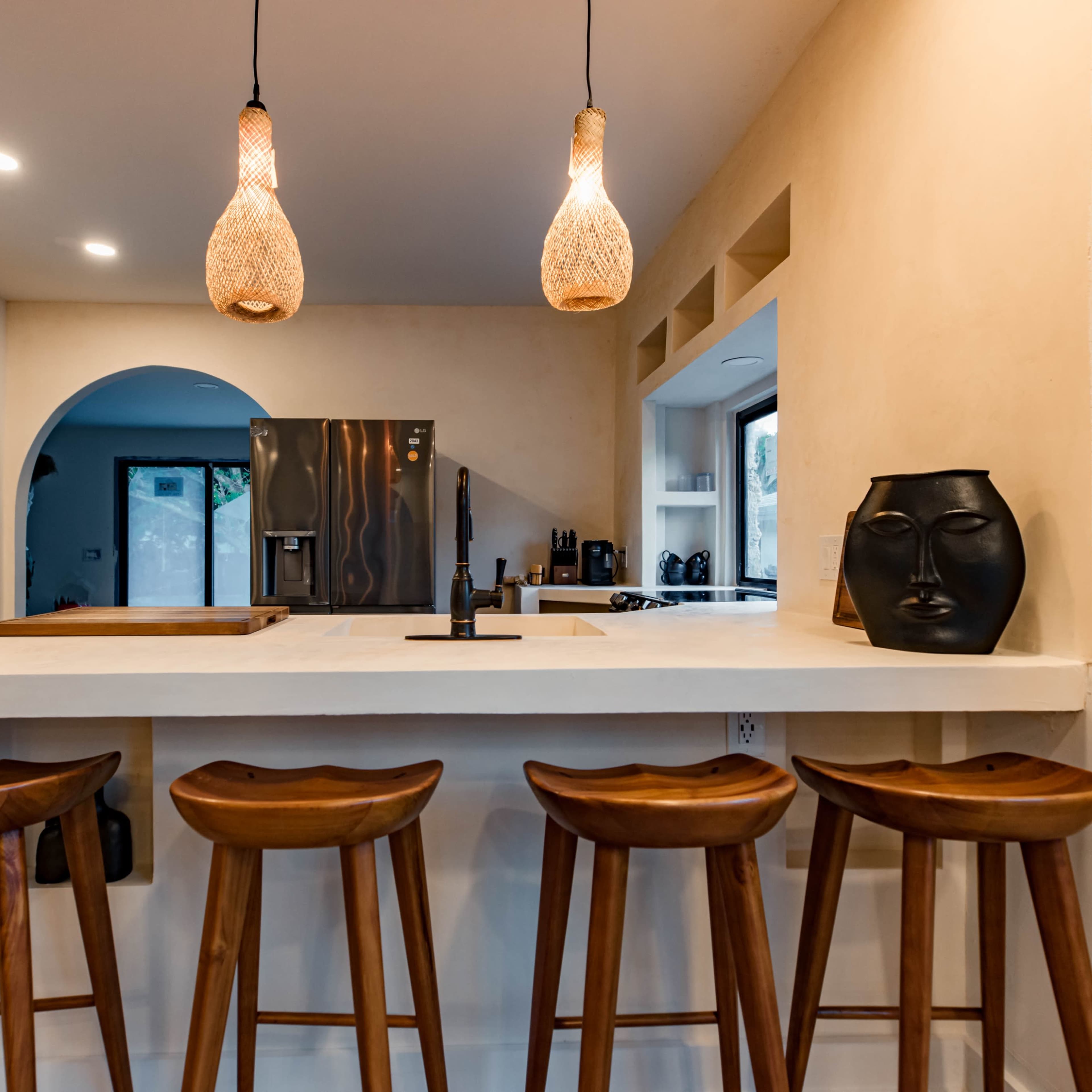 A modern kitchen with a white countertop, wooden stools, and pendant lighting above, featuring a doorway that leads to another room.