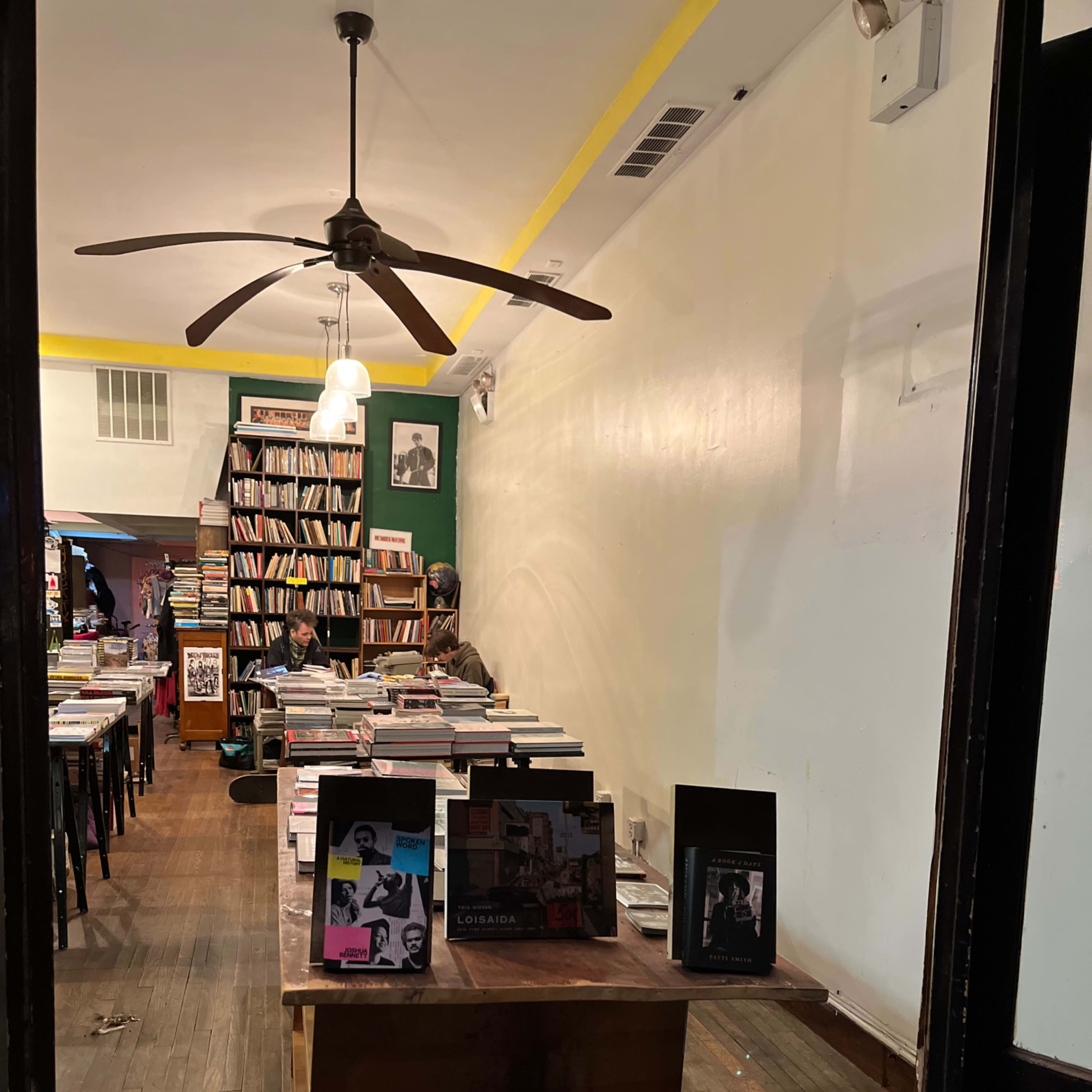 The interior of a bookstore with wooden floors, a long table displaying books, and a bookshelf filled with various titles in the background.