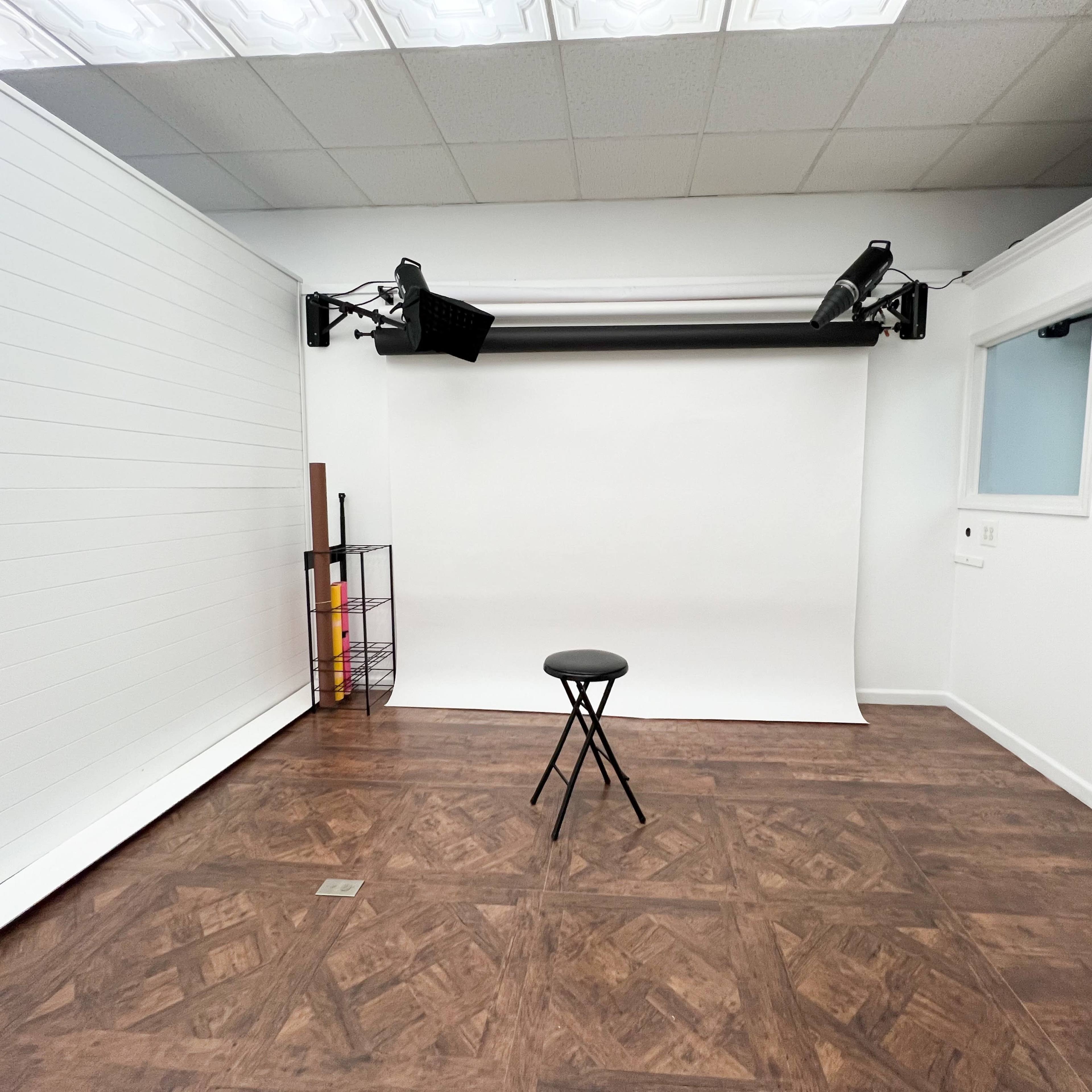 A photography studio featuring a simple setup with a plain white backdrop, a black stool in the center, and minimal equipment mounted on the ceiling.