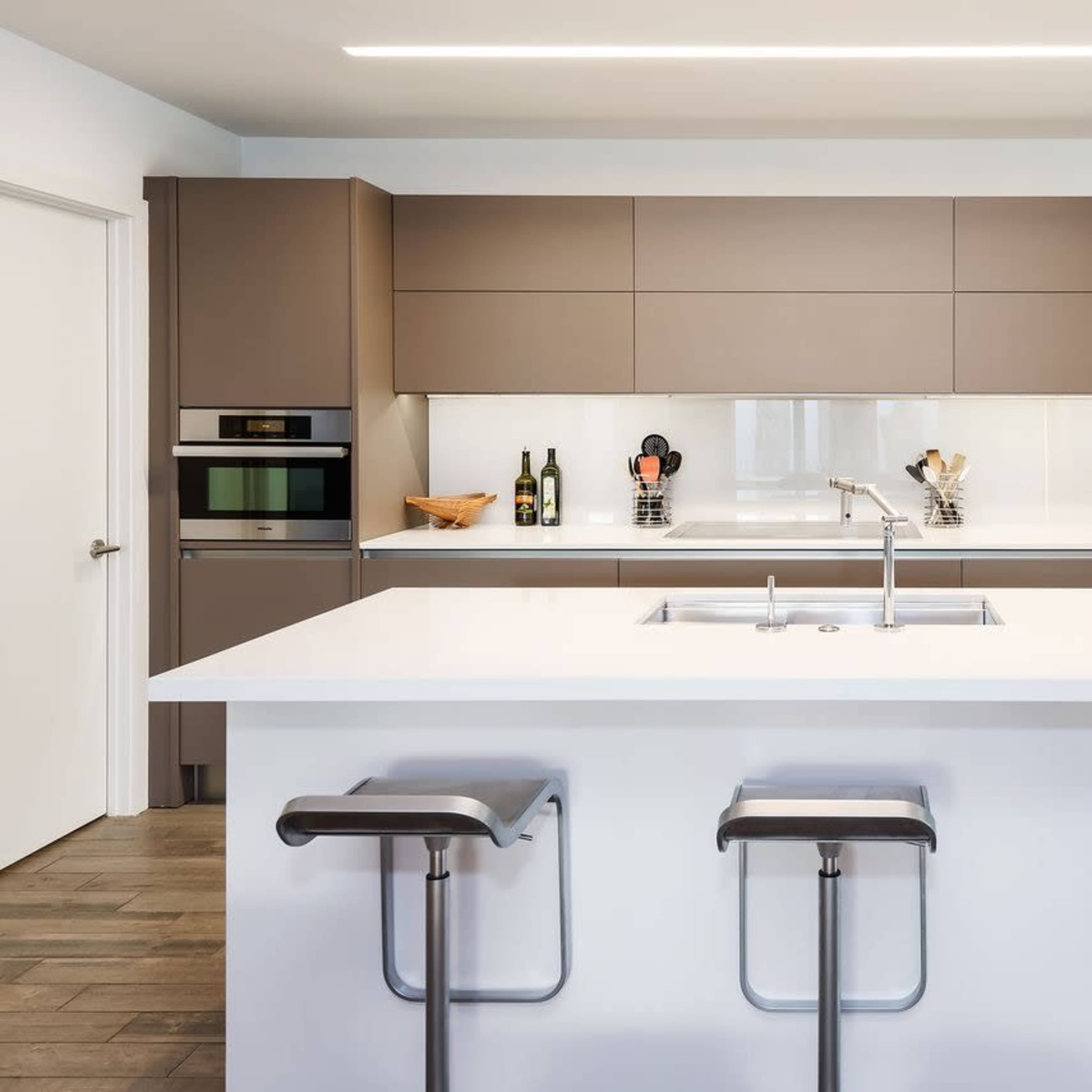A modern kitchen features a central island with bar stools, sleek cabinetry, and built-in appliances against a light-colored wall.