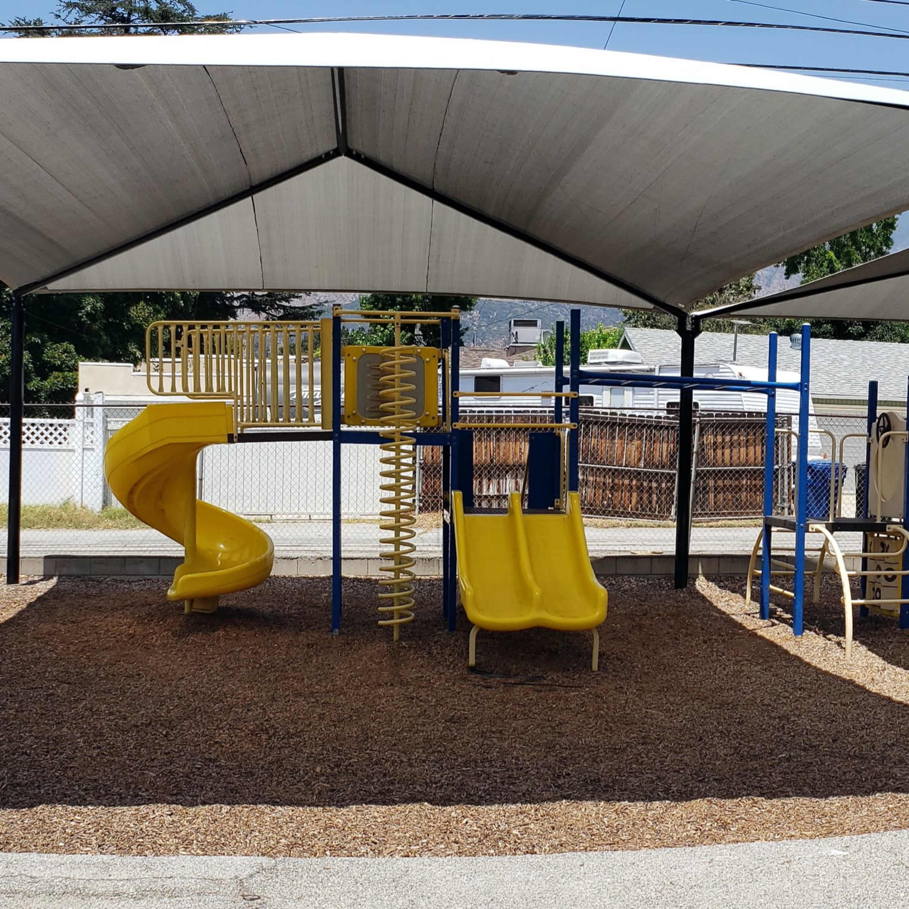 A playground area with yellow slides, a small playhouse, and shaded structures surrounded by a gravel surface.