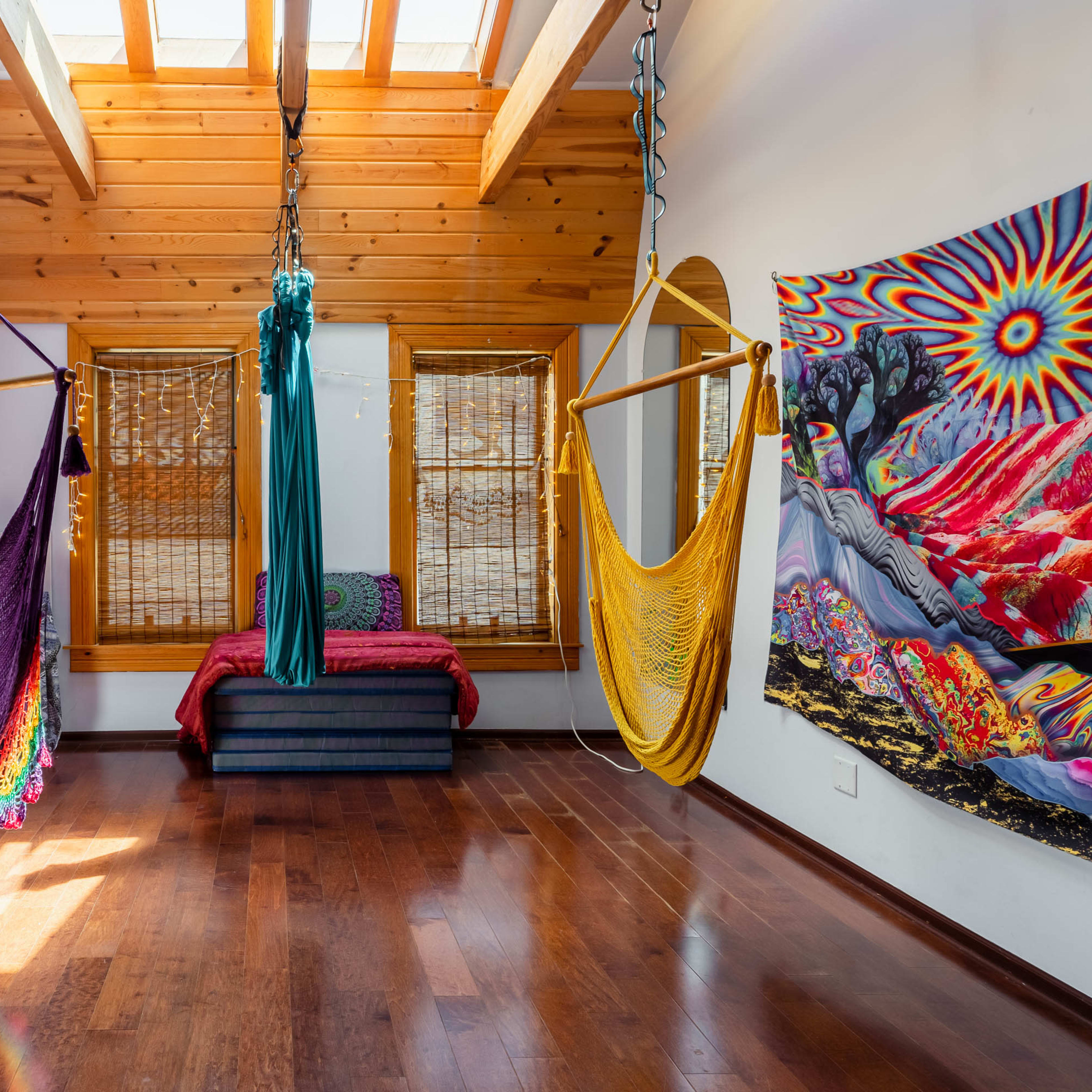 A room with wooden beams and a skylight, featuring colorful hammocks and a large tapestry on the wall, alongside a mirrored surface and rustic furnishings.