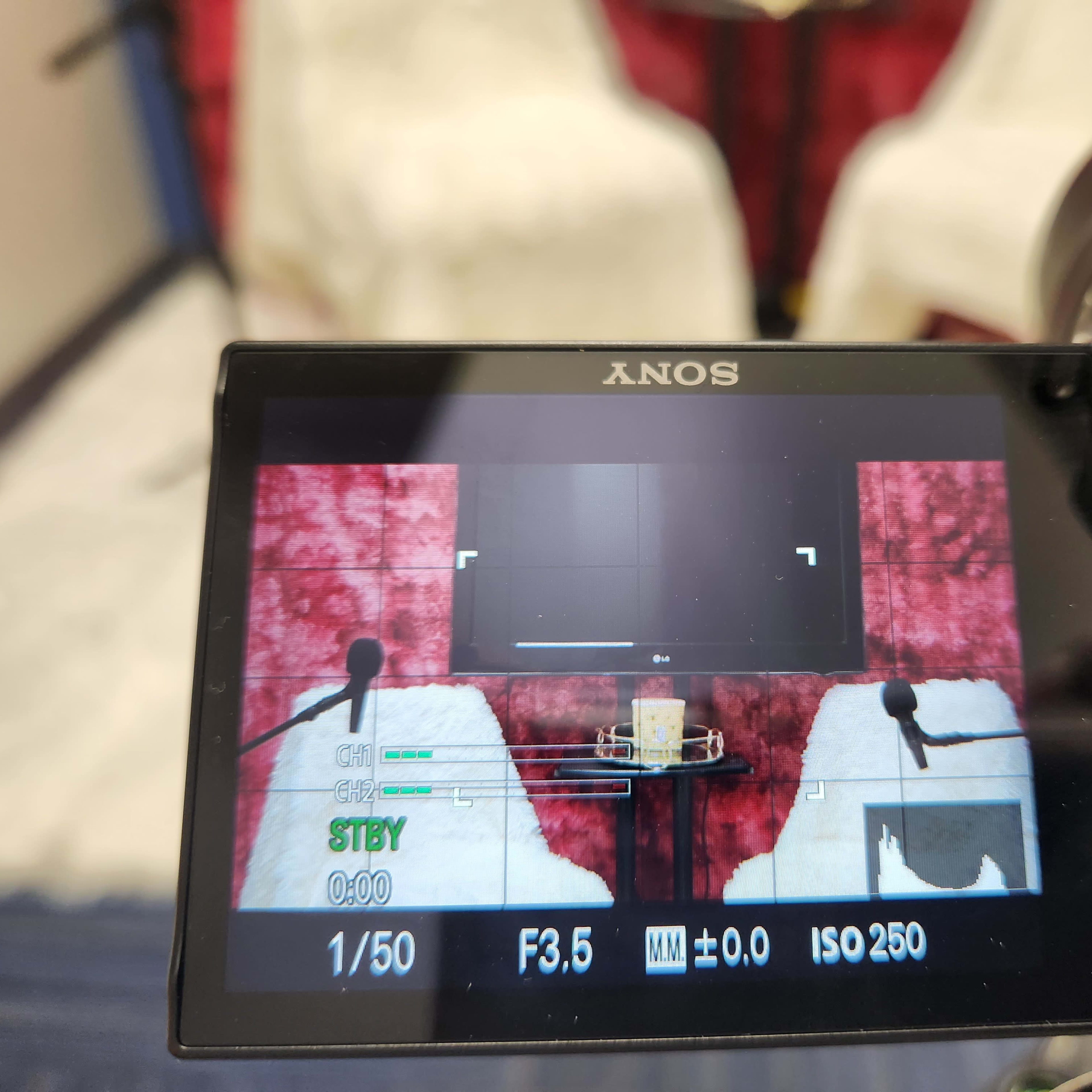 The camera viewfinder shows a setup with two microphones in front of empty white chairs and a screen covered in a red backdrop.