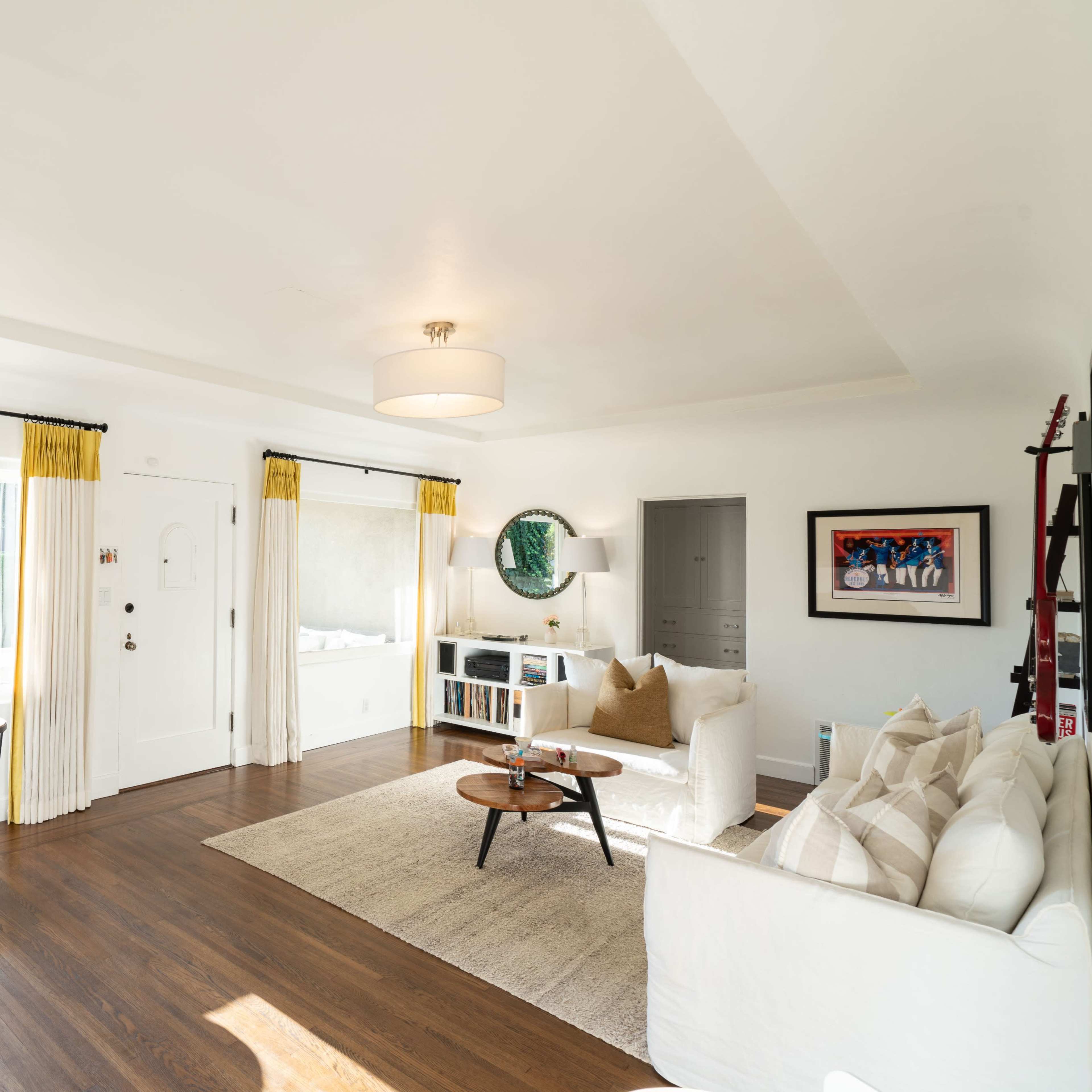 A well-lit living room features two white sofas, a round coffee table, and large windows with yellow curtains.