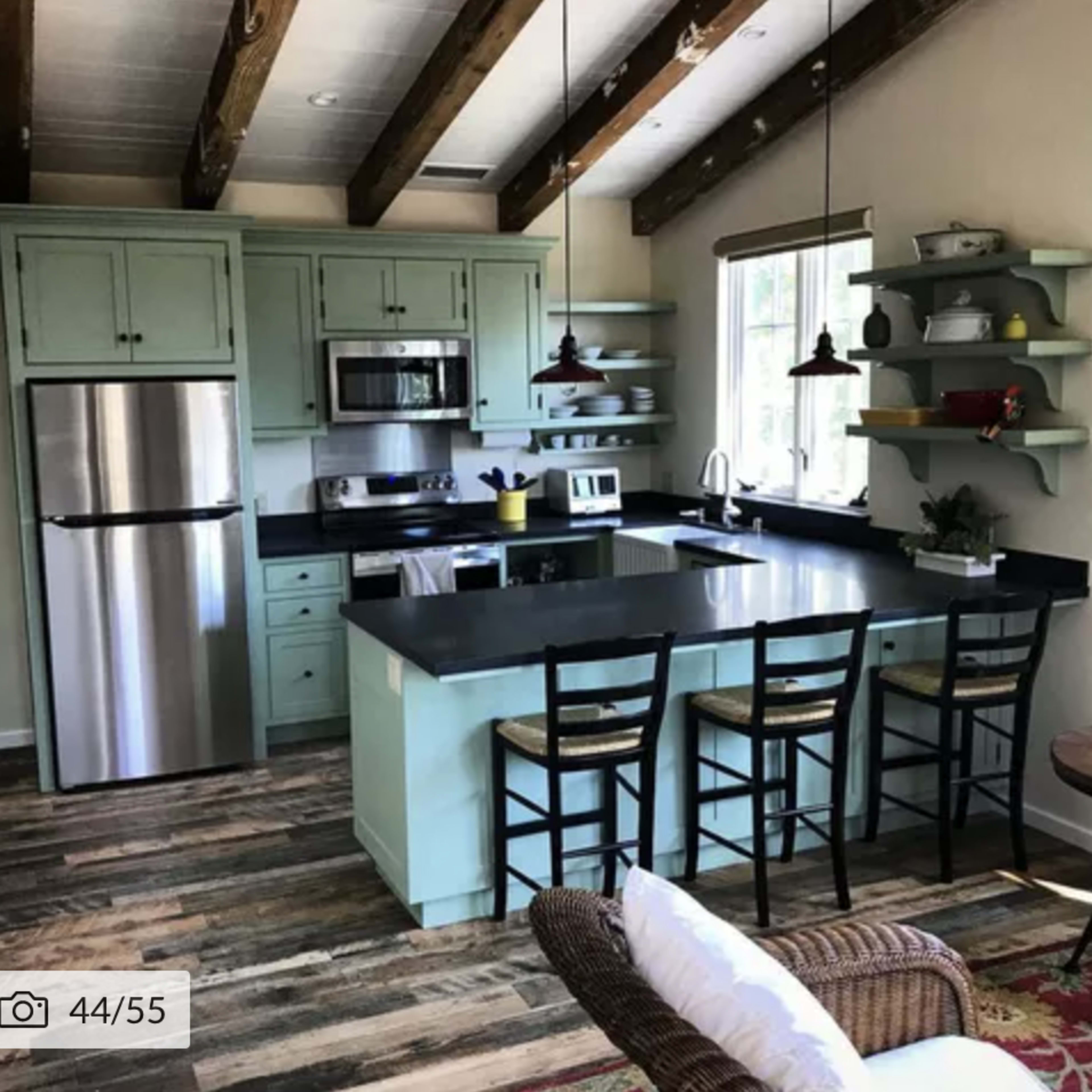 A modern kitchen with mint green cabinets, stainless steel appliances, and a black countertop, featuring an island with four chairs and wooden beams on the ceiling.